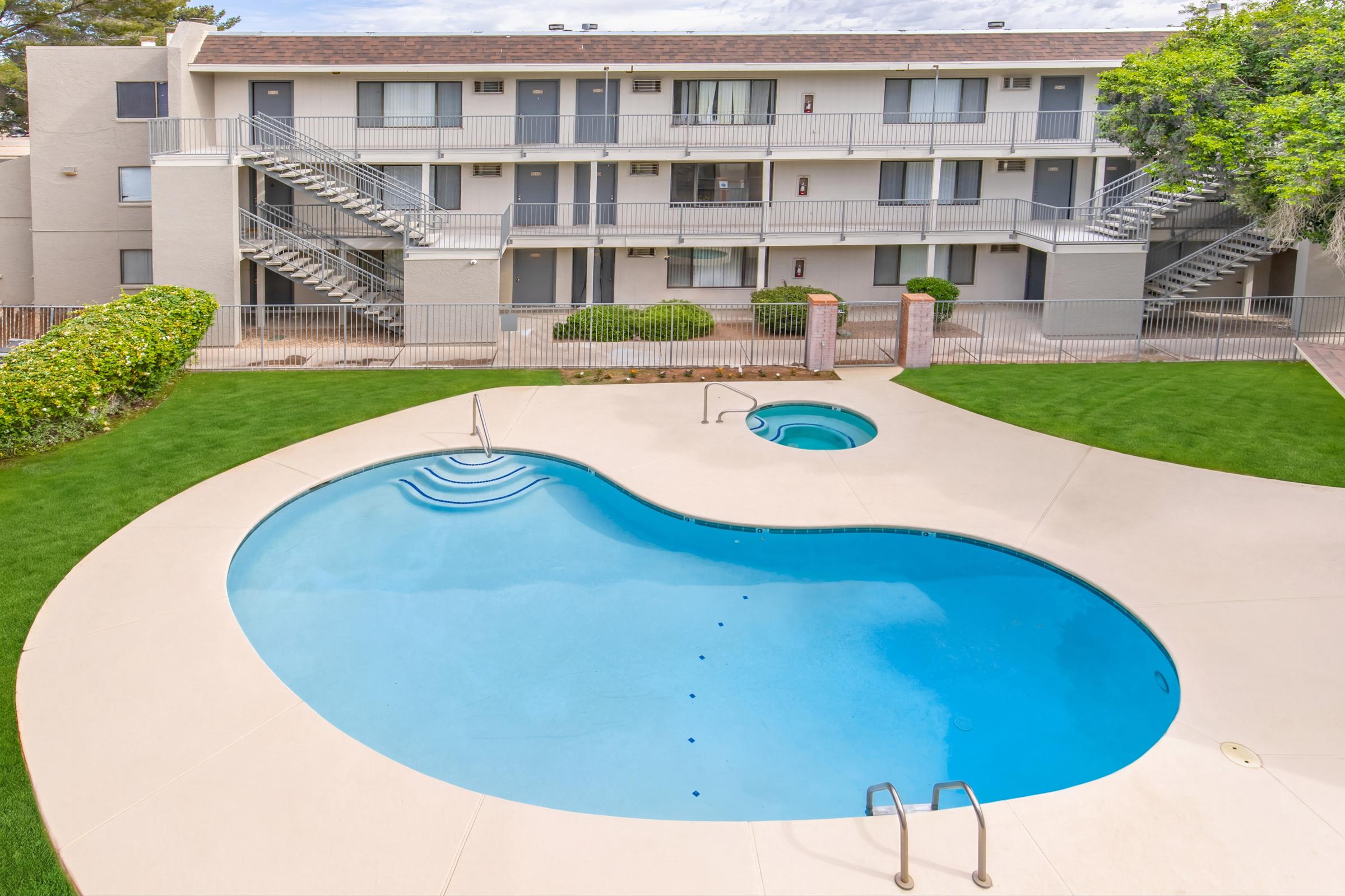 A view of a multi-story apartment complex featuring a swimming pool and a smaller hot tub. The pool is shaped organically and surrounded by a well-maintained green lawn. Staircases lead to the upper floors of the building, with several balconies visible. The sky is partly cloudy in the background.