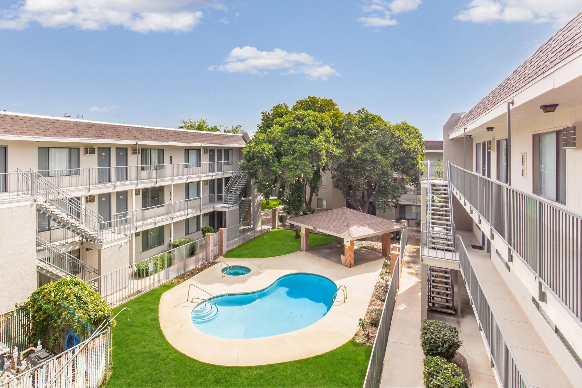 A view of a hotel courtyard featuring a small swimming pool surrounded by grass, with trees providing shade. The image shows two multi-story buildings with balconies overlooking the pool area, and a gazebo nearby, all set against a clear blue sky.