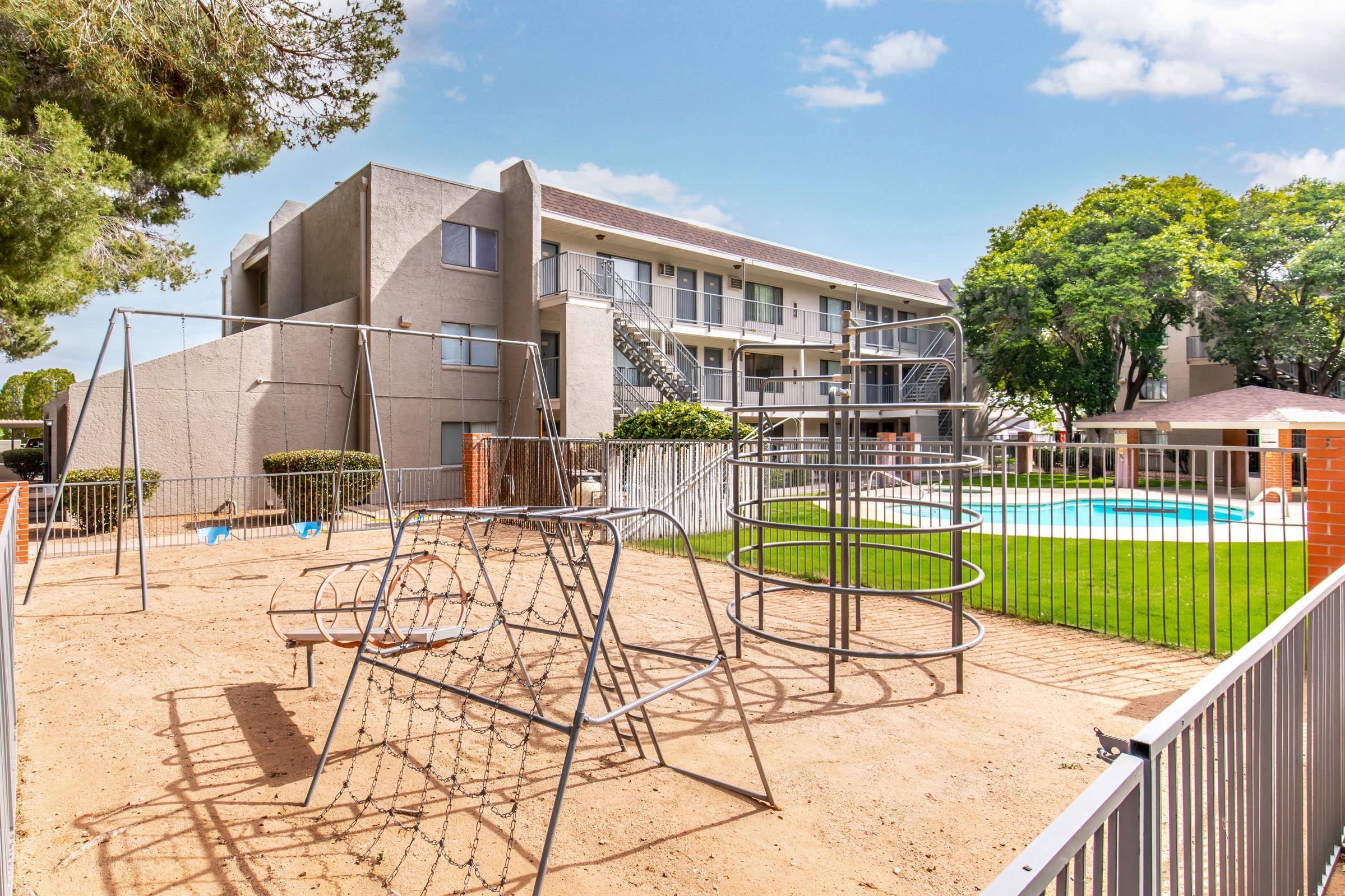 A playground area featuring swings and a climbing structure, enclosed by a fence. In the background, there is an apartment building with a pool surrounded by green lawn and trees under a blue sky.
