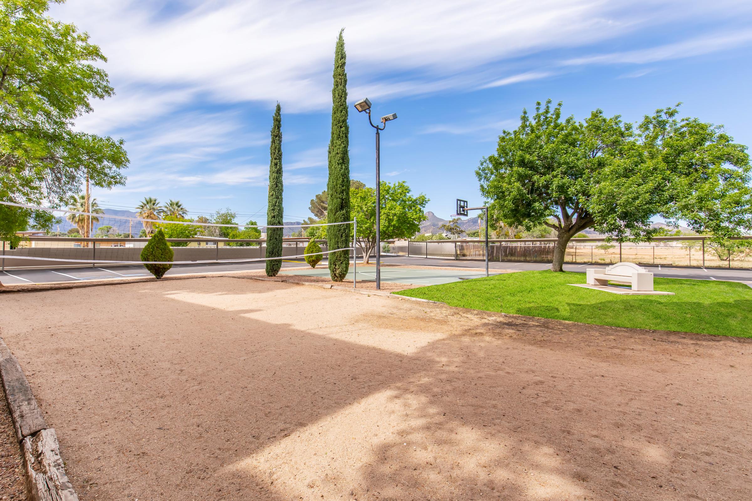 A spacious outdoor area featuring a sand volleyball court on the left, lined with two tall trees. In the background, there's a basketball court and green grass. A few palm trees are visible, along with distant mountains. The sky is blue with some white clouds, creating a vibrant and inviting atmosphere.