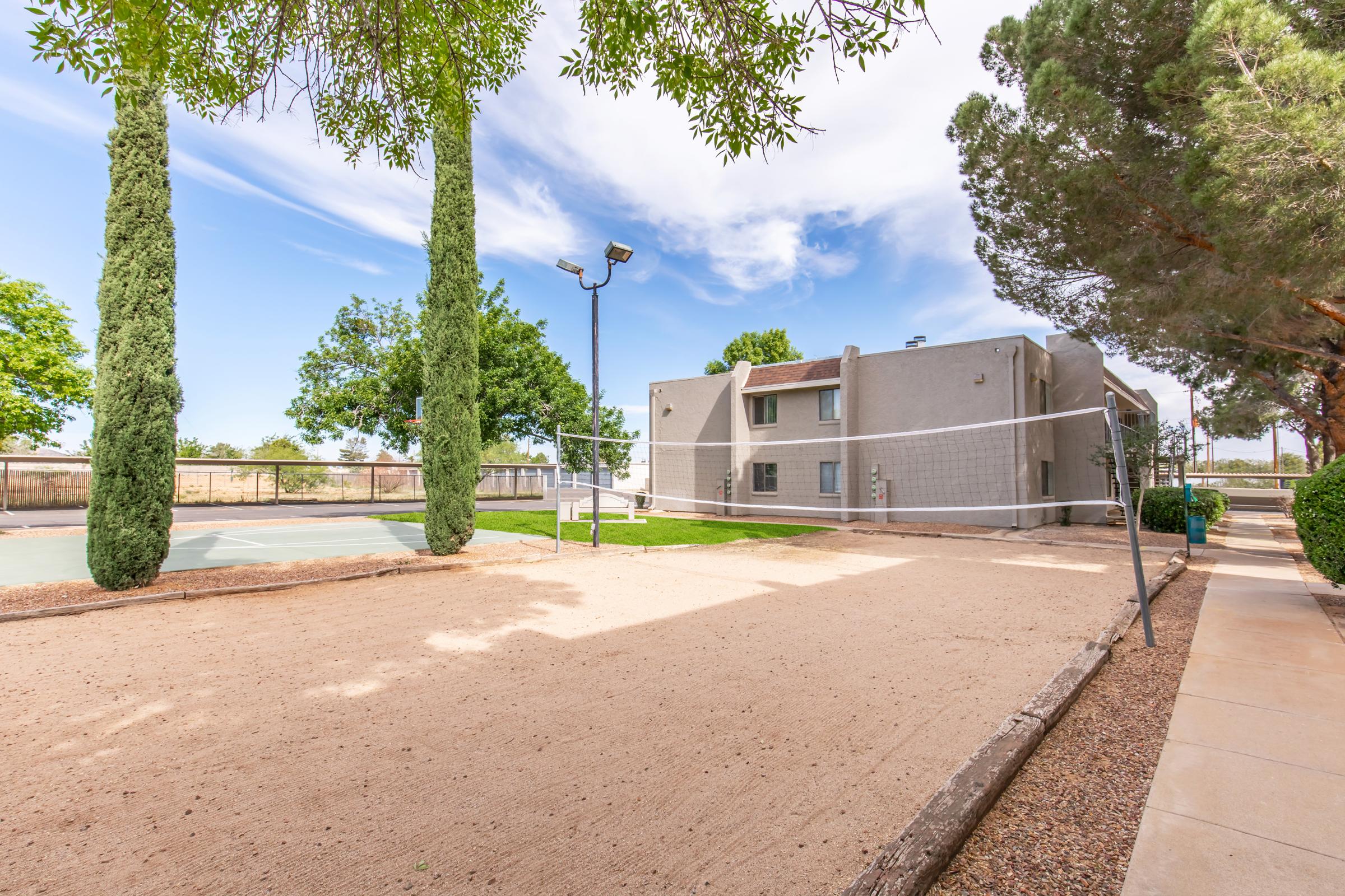 A sandy volleyball court surrounded by greenery, with tall trees and a building in the background. The sky is bright blue with a few clouds. Floodlights are present, indicating the area can be used for evening games. A well-maintained landscape complements the recreational space.