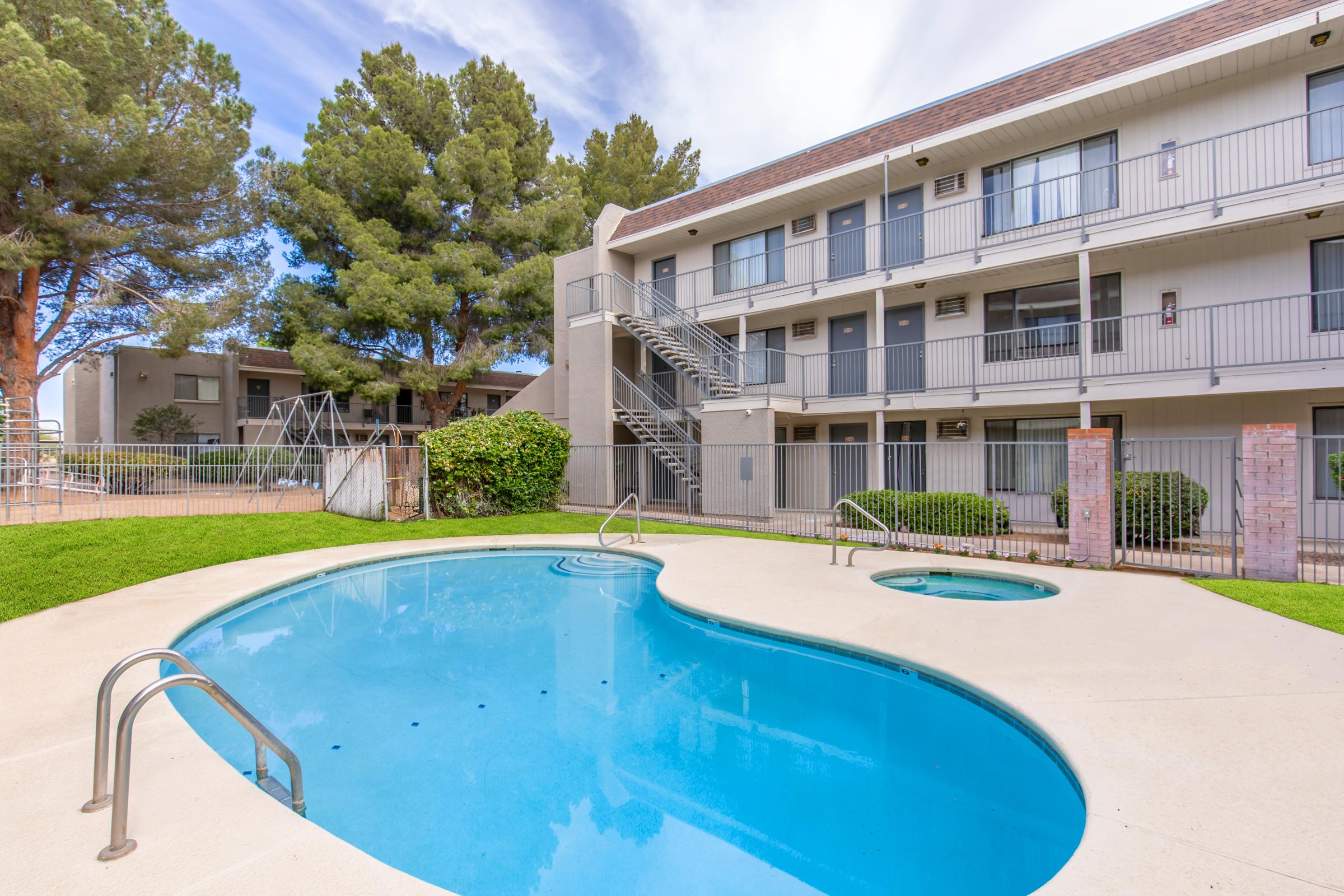 A clear, inviting swimming pool with a hot tub, surrounded by lush green grass and tall trees. In the background, a three-story apartment building with balconies can be seen, along with a playground area. The sky is partly cloudy, creating a pleasant outdoor atmosphere.
