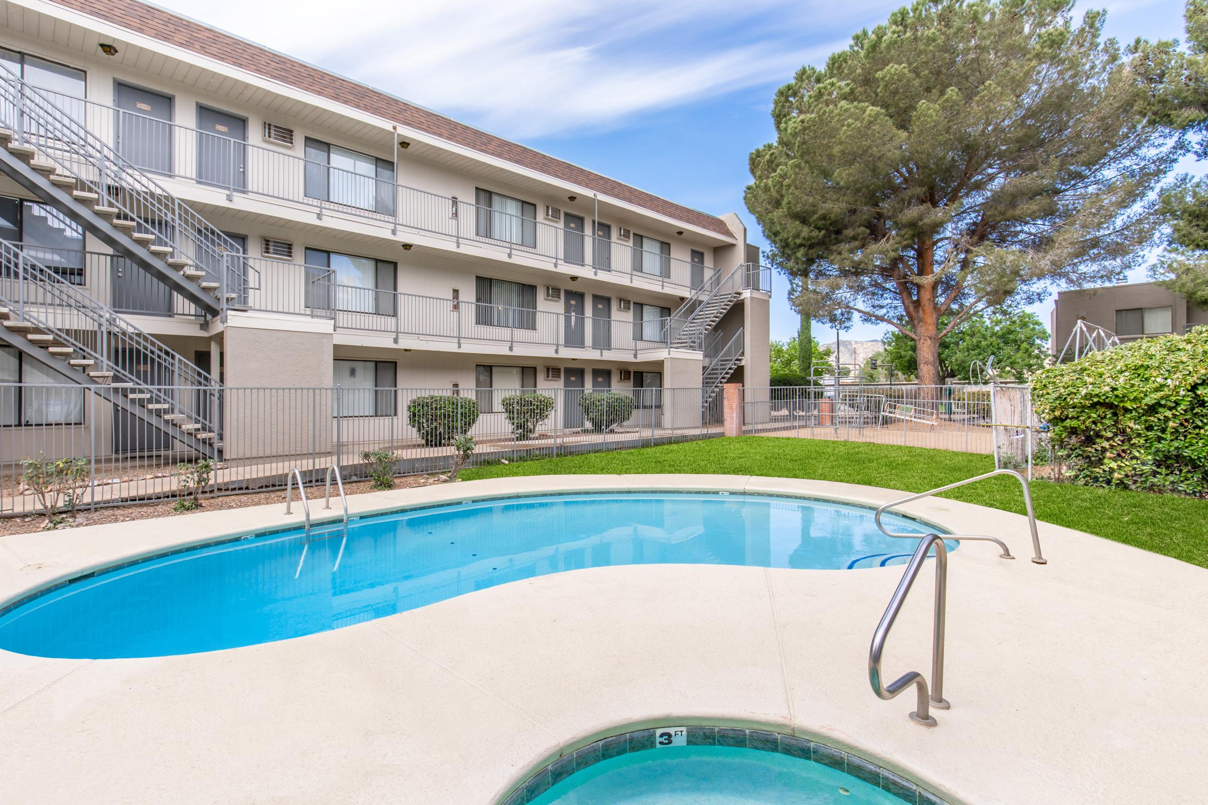 A swimming pool with a hot tub, surrounded by grass and shrubs, at an apartment complex. The building features multiple levels with staircases. Sunny sky and trees nearby create a relaxing outdoor atmosphere.