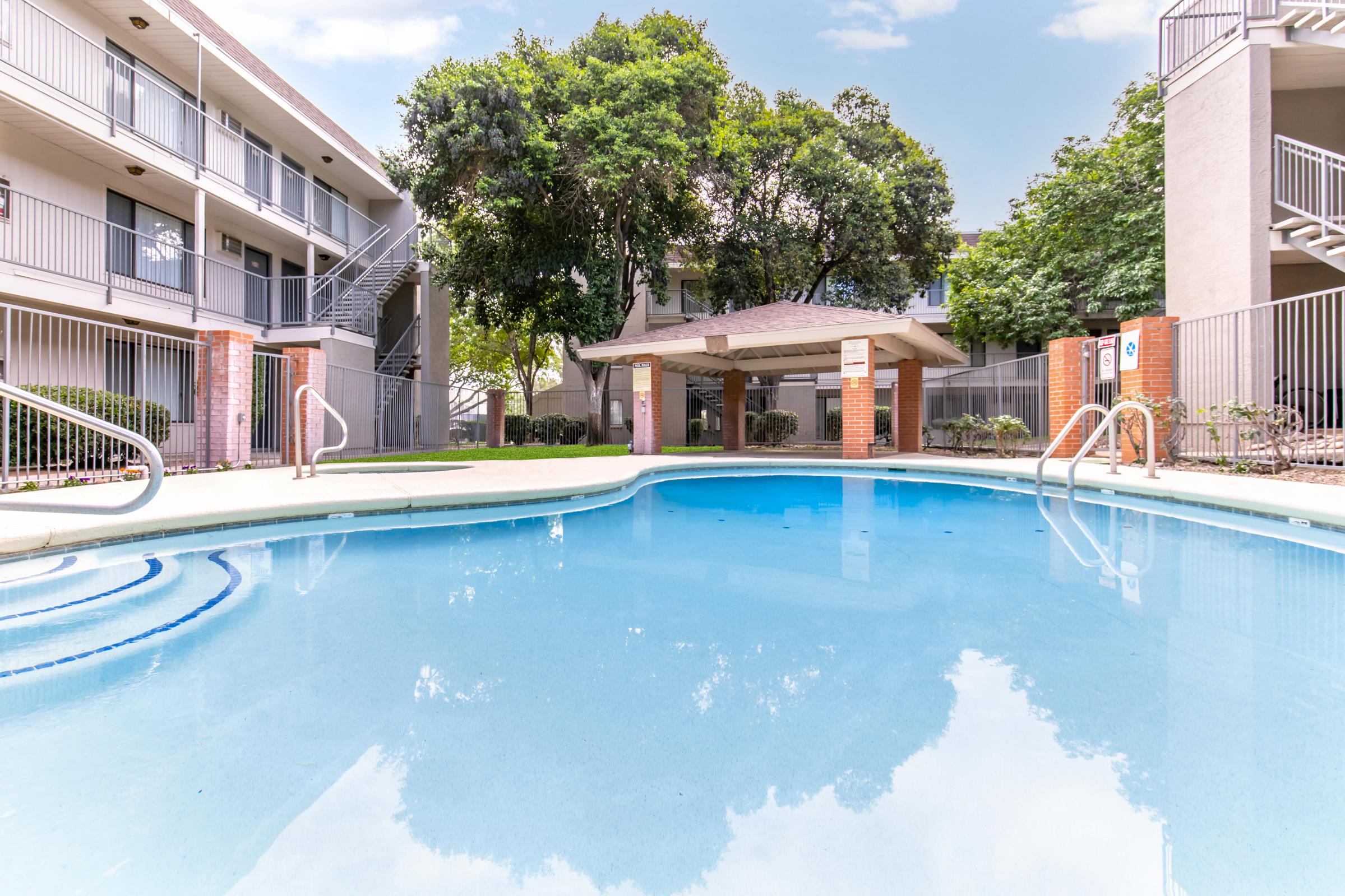 A clear blue swimming pool surrounded by lush greenery and apartment buildings. A shaded gazebo is visible nearby, with chairs and tables arranged around the pool area. The sky reflects in the water, creating a serene atmosphere. Steps lead into the pool from the deck.