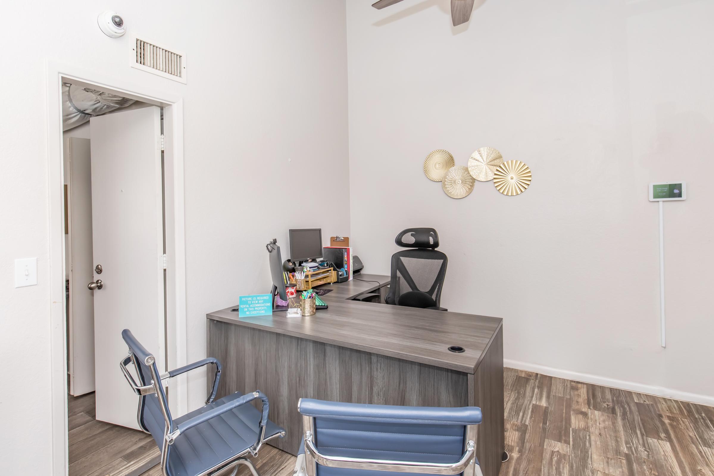 An office space featuring a modern desk with a computer, office supplies, and a stylish chair. Two blue chairs are positioned in front of the desk. The walls are light-colored, adorned with decorative circular wall art, and a thermostat is visible on one wall. There's a door leading outside of the room.
