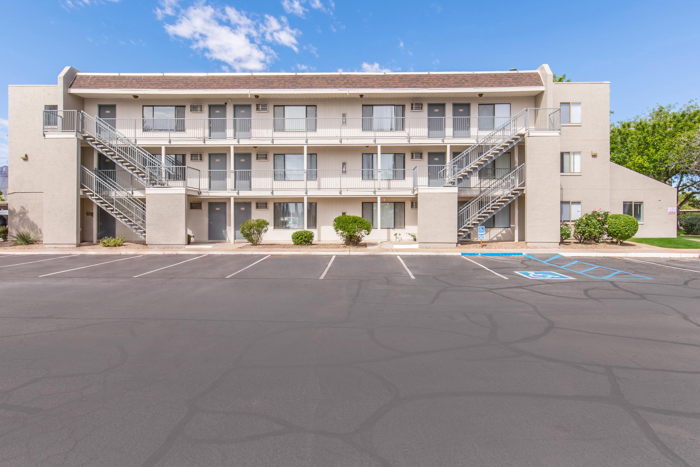 A two-story apartment building featuring a light-colored exterior with multiple balconies. Outdoor stairs lead to upper units. The surrounding area includes small shrubs, and a paved parking lot with visible handicap spaces. The sky is clear with a few clouds.