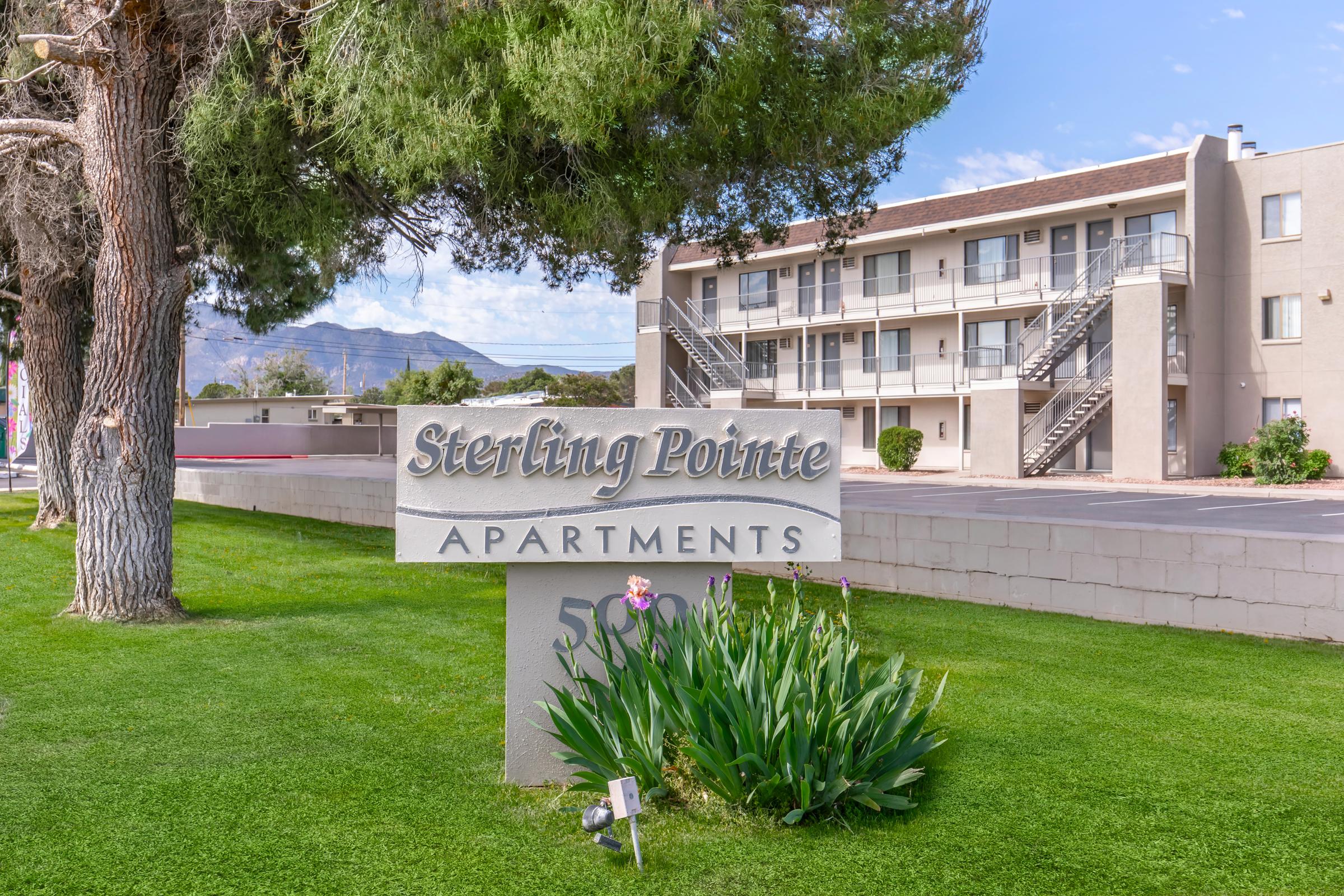 Sign for Sterling Pointe Apartments in front of a multi-story building with a green lawn and trees. The building features balconies and outdoor staircases. The background includes mountains and a clear blue sky.