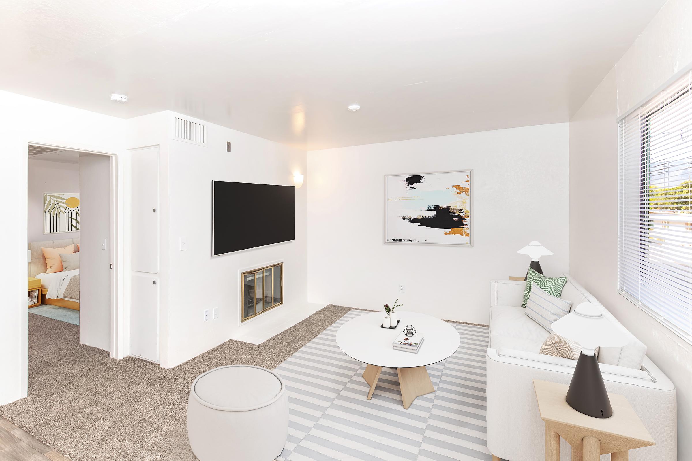 A bright, modern living room featuring a white sofa with colorful cushions, a round coffee table on a striped rug, and a wall-mounted flat-screen TV. Light floods the space through a window, complemented by a minimalist abstract artwork on the wall. A cozy atmosphere with neutral tones and soft textures.