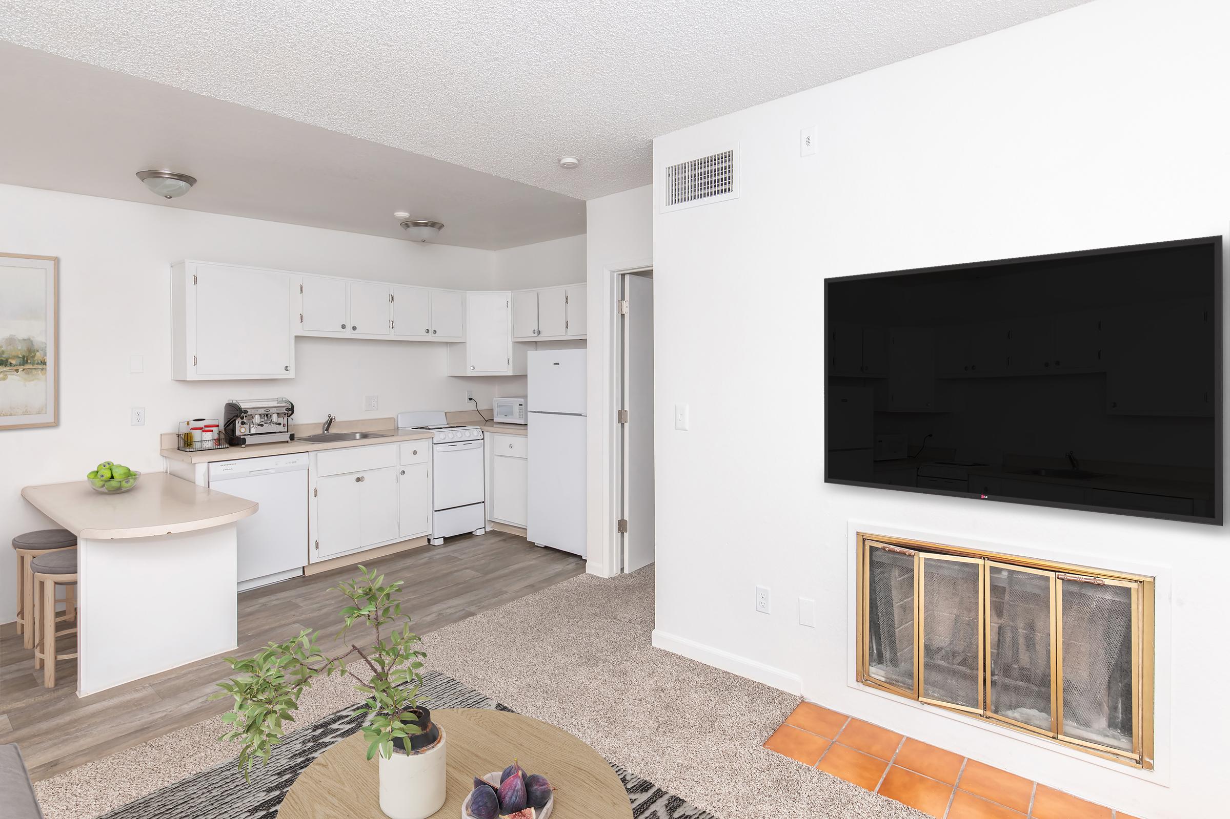 A modern, bright kitchen and living area featuring white cabinetry, a refrigerator, and an oven. There’s a comfortable seating area with a round table and a potted plant. A large, wall-mounted flat-screen TV is on one wall, and a decorative fireplace is nearby. The flooring is light wood with carpets in the living area.