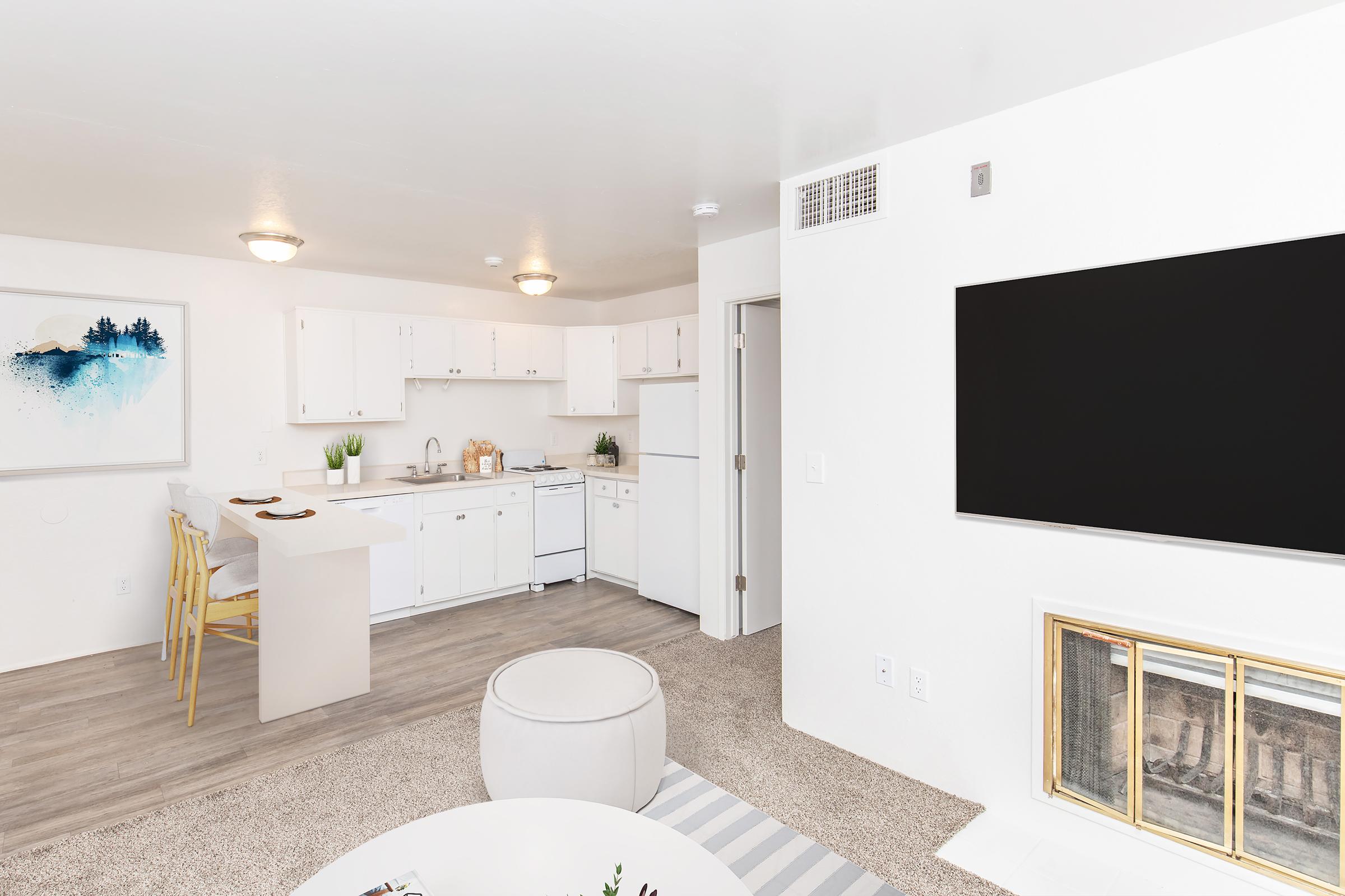 A bright, modern living space featuring an open kitchen with white cabinetry, a stove, and a refrigerator. There's a cozy seating area with a round ottoman and a large wall-mounted TV. The room has light-colored walls, neutral carpeting, and a few decorative plants, creating a welcoming atmosphere.
