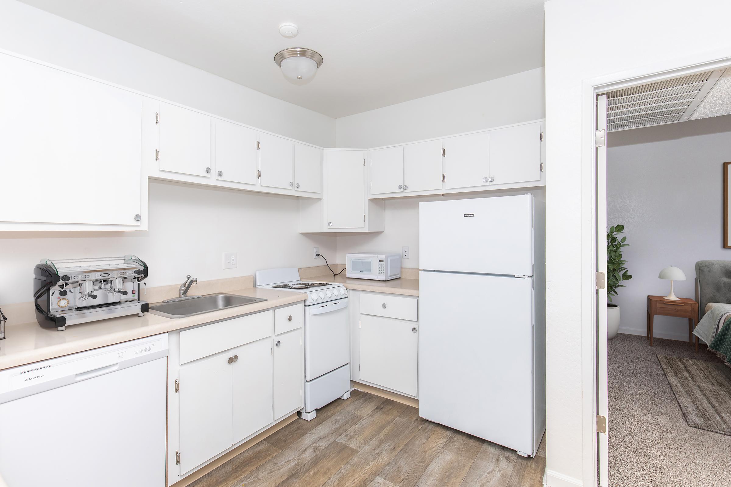 A bright and clean kitchen featuring white cabinets, a sink, a stove, a microwave, and a refrigerator. The countertop is clutter-free, and the flooring has a wood-look design. A doorway leads to a living area in the background.