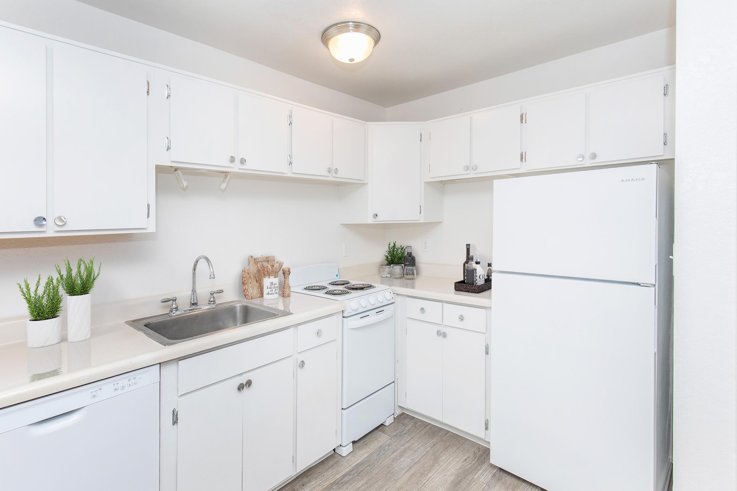 A modern, bright kitchen featuring white cabinets, a white refrigerator, a stove, and a sink. The countertops are light-colored, and there are decorative plants on the counter. The space is tidy and well-organized, with natural light illuminating the room.