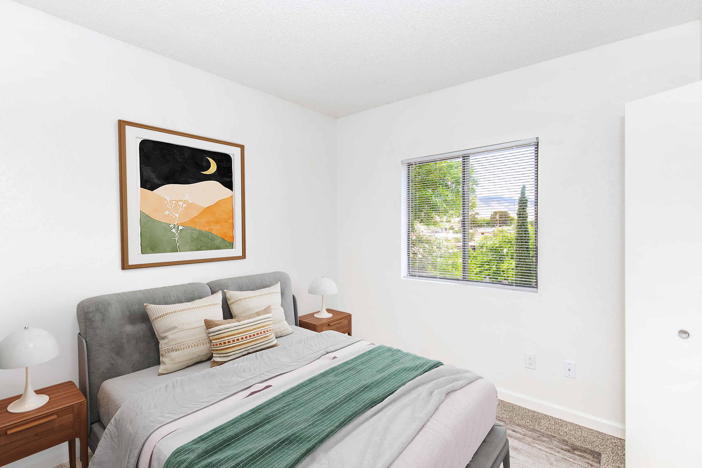 A cozy bedroom with a gray upholstered bed featuring decorative pillows and a green throw. On either side of the bed are wooden nightstands with white lamps. A large framed abstract artwork is on the wall, and a window offers a view of greenery outside, providing natural light. The walls are painted white, enhancing the bright atmosphere.