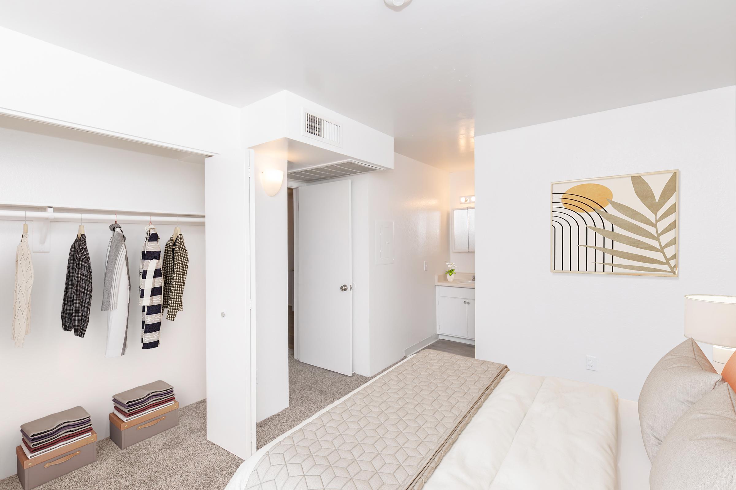 A bright, modern bedroom with a neatly made bed and a light beige quilt. To the left, a closet with hanging clothes is visible, while the right side features a bathroom door. A framed abstract artwork hangs on the wall, and storage boxes are placed at the foot of the bed, all set against a clean, white interior.