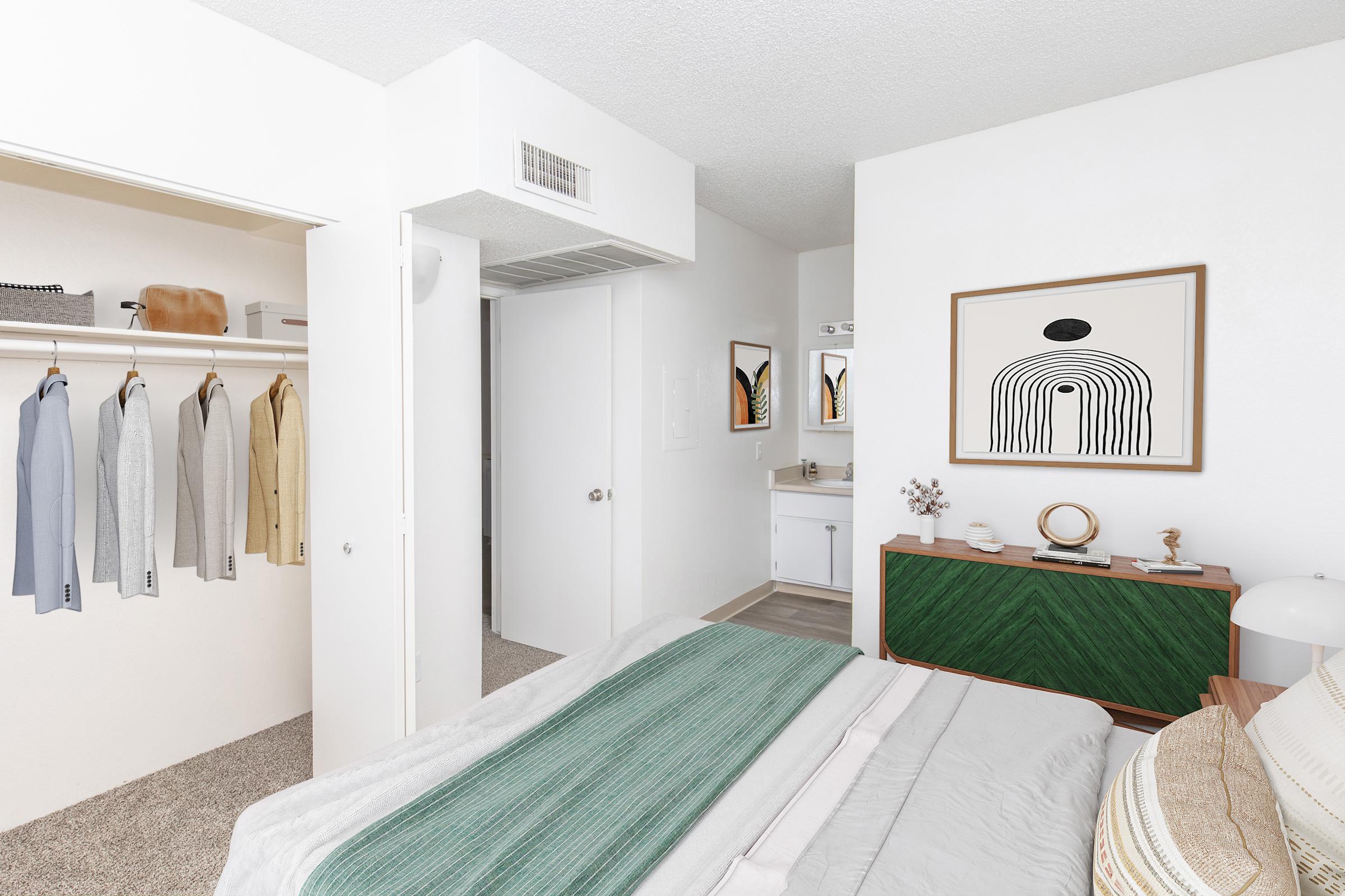 A modern bedroom with a neatly made bed featuring a green and gray blanket. To the left, there's a closet with various hanging clothes. The room has a white color scheme, with artwork on the walls and a green dresser. There's a small vanity area with mirrors, adding to the stylish decor.