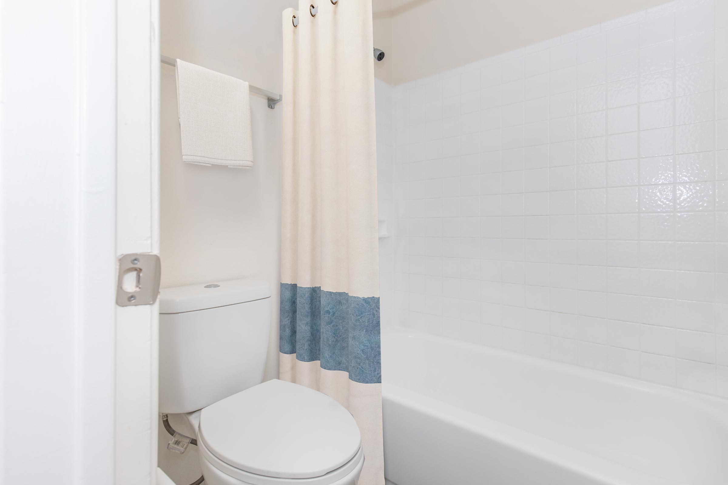 A clean bathroom featuring a white bathtub, a toilet, and a shower with a beige curtain that has a blue stripe. White tiled walls and a roll of towels are visible on a rack above the toilet. The space is well-lit and organized.