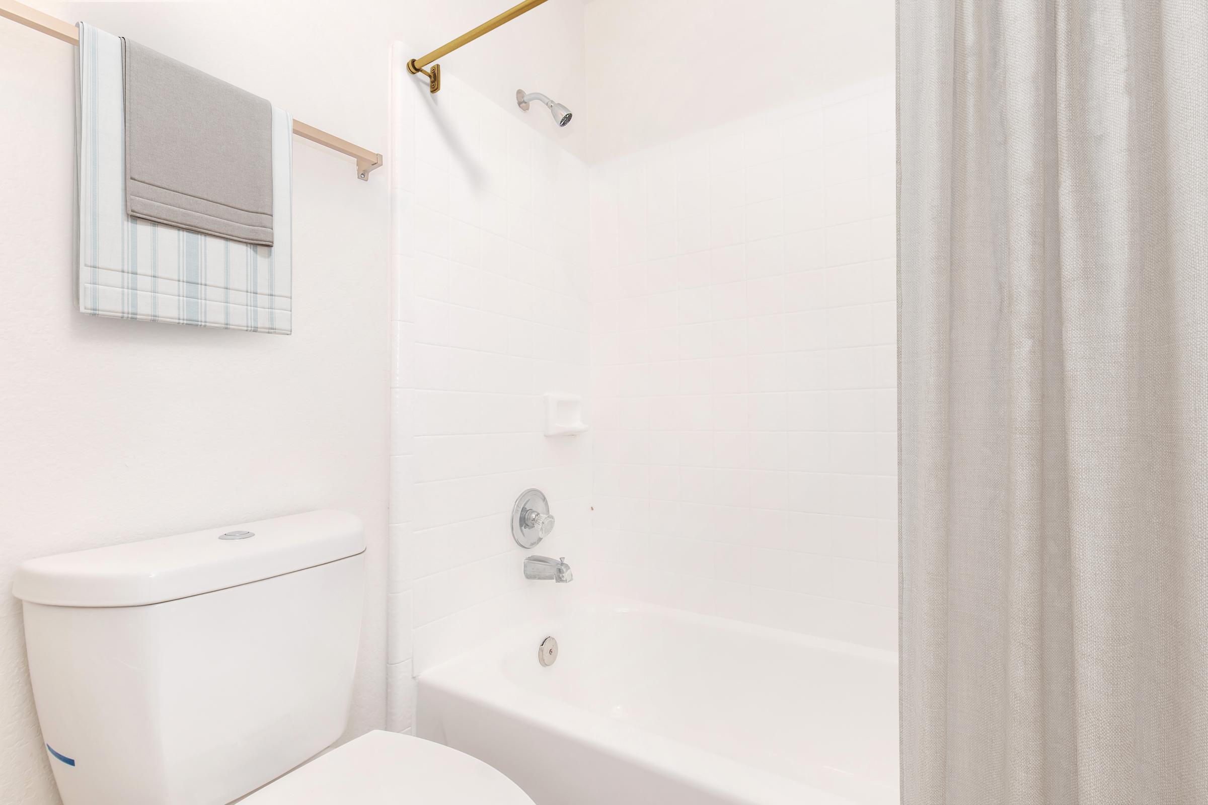 A clean bathroom setup featuring a white bathtub, a shower with a curtain, a modern toilet, and neatly hung towels on a rack. The walls are painted in a light color, contributing to a fresh and airy atmosphere. The space is well-organized and minimalistic.
