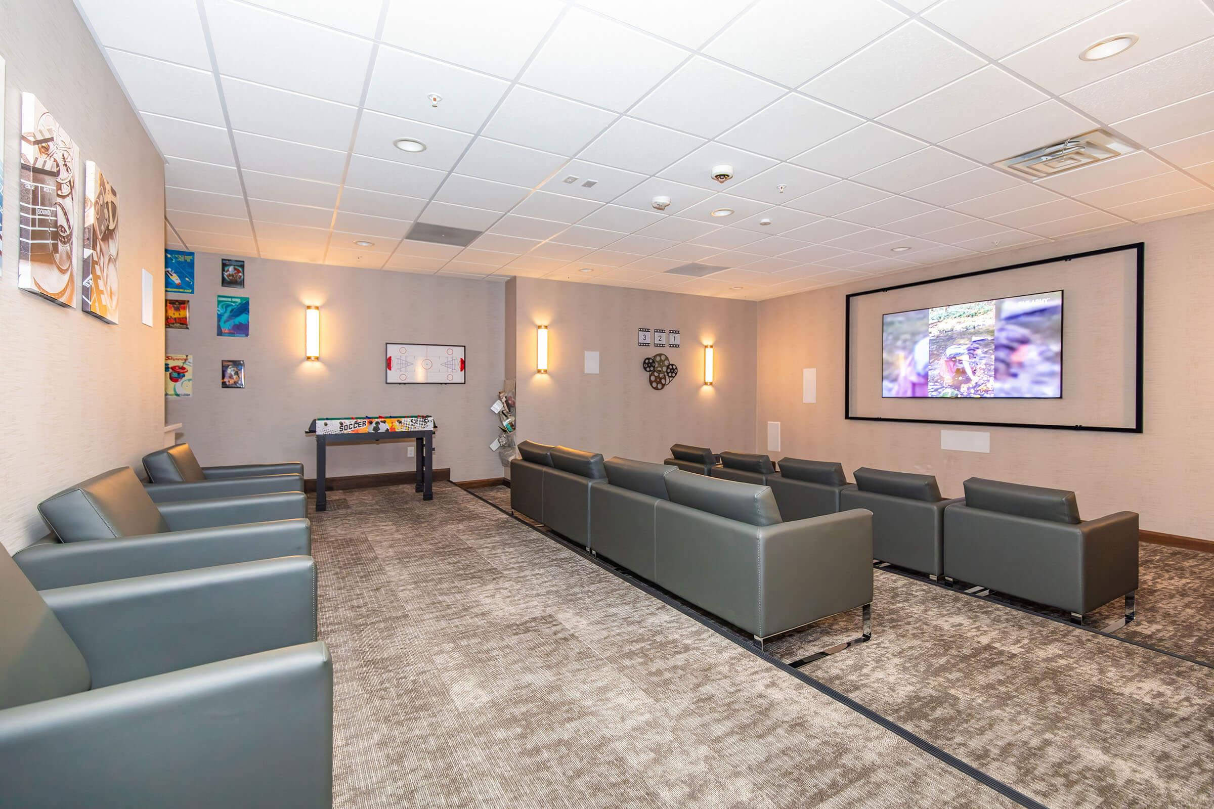 A modern waiting room featuring gray leather seating, a large screen displaying a video, and a wall decorated with various images and artworks. The space is well-lit with soft lighting and has a carpeted floor, creating a comfortable and inviting atmosphere.