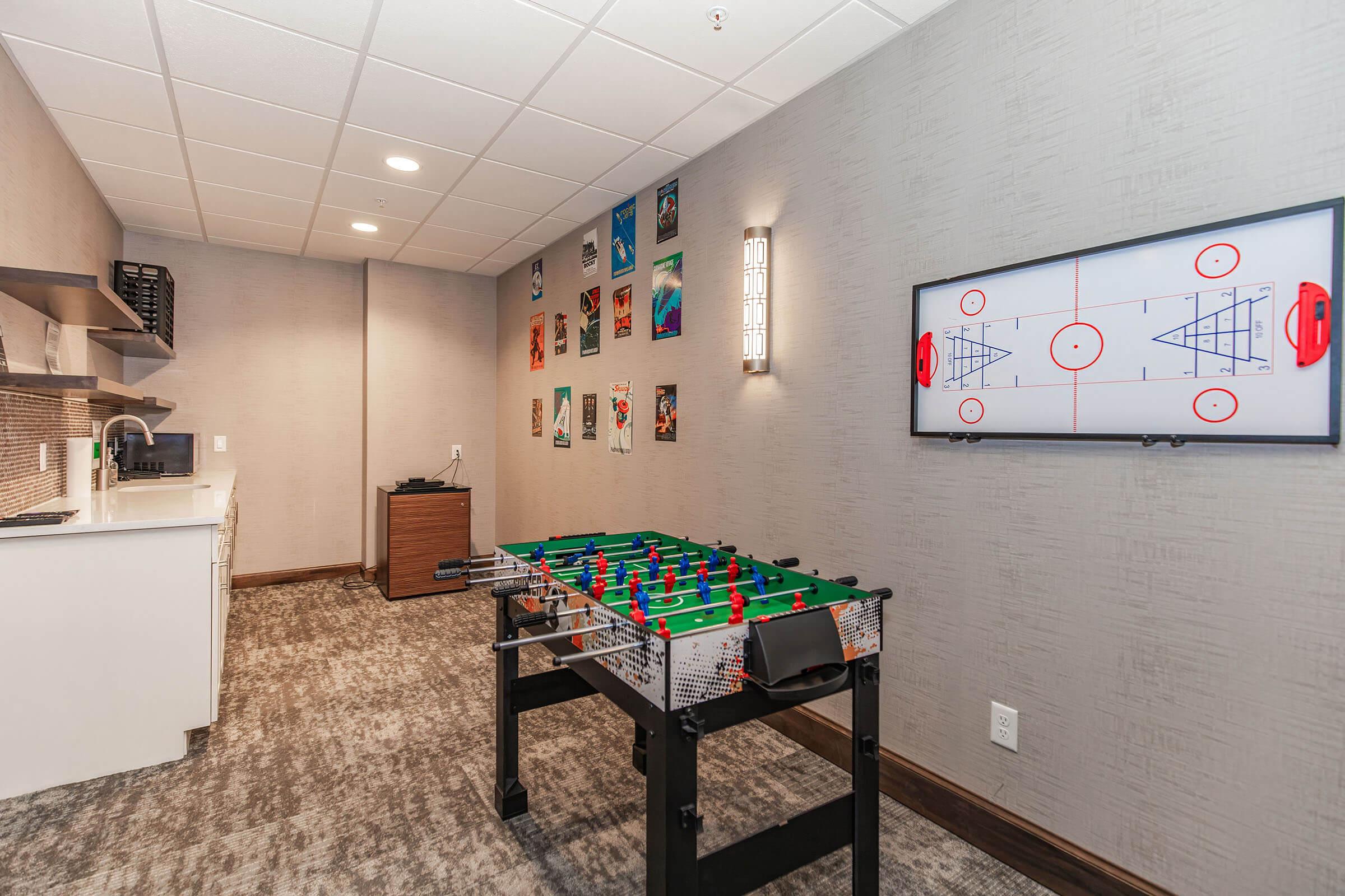 A recreational room featuring a foosball table, a wall-mounted television displaying a sports layout, and framed sports posters on the gray walls. A kitchenette with cabinets is visible in the background, and the floor is carpeted. The lighting is bright, creating an inviting atmosphere for entertainment and relaxation.