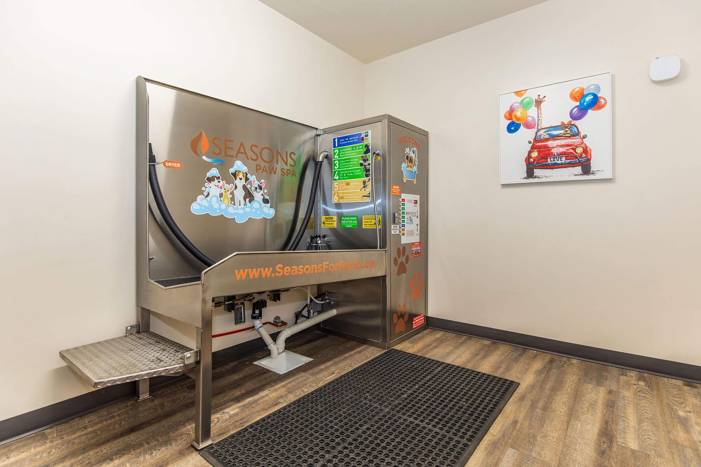 Self-service pet washing station featuring a bright, metallic design with colorful graphics of dogs. The machine includes controls for settings and a small platform for pets. Nearby, a wall displays balloons and a car, enhancing the playful atmosphere. The floor is made of dark wood-like material.