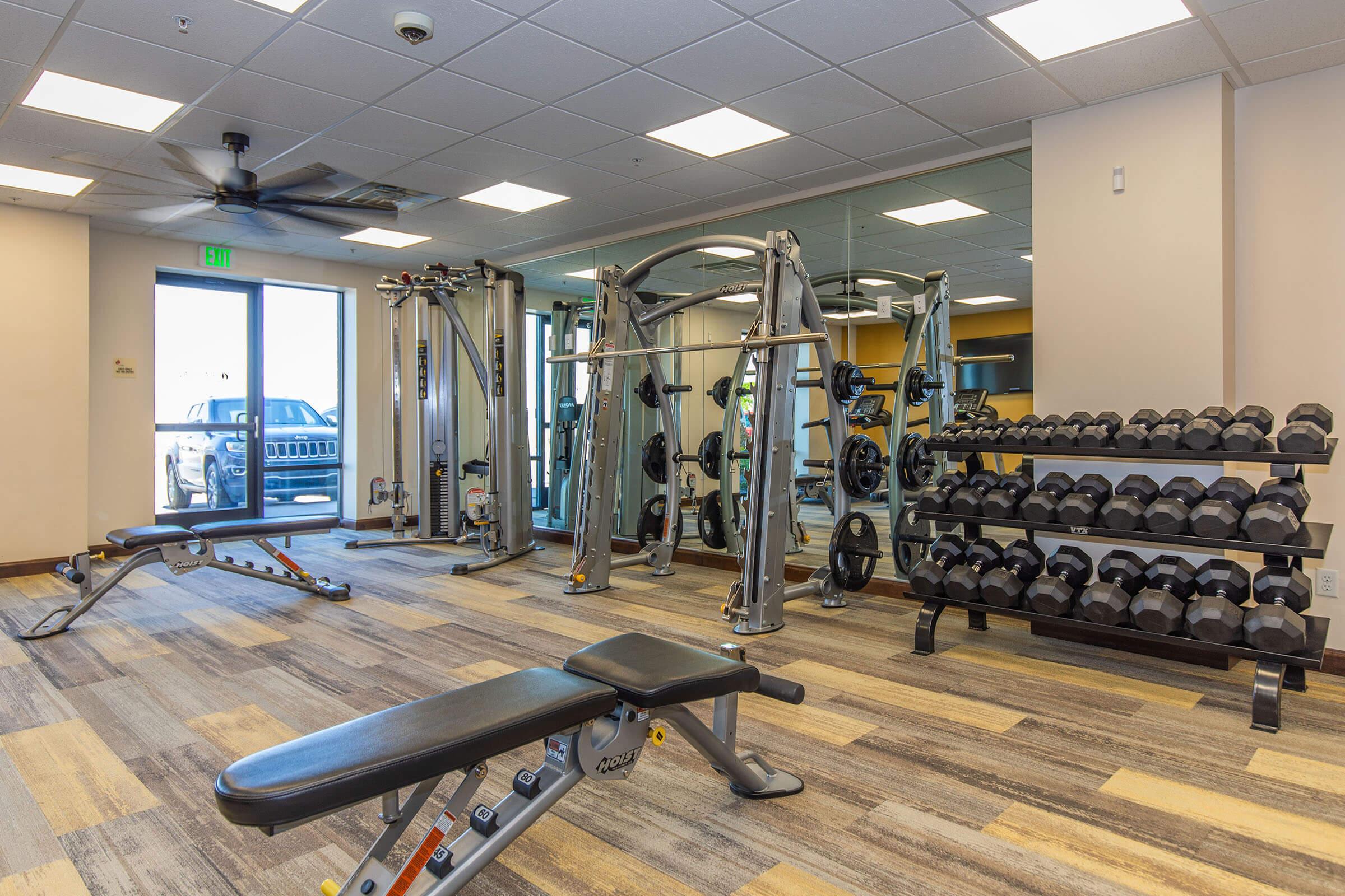 A well-equipped gym featuring weight machines, free weights on a rack, and a bench for exercises. Large mirrors and windows create an open atmosphere, while a ceiling fan adds ventilation. The space is bright and modern, suitable for a variety of fitness workouts.