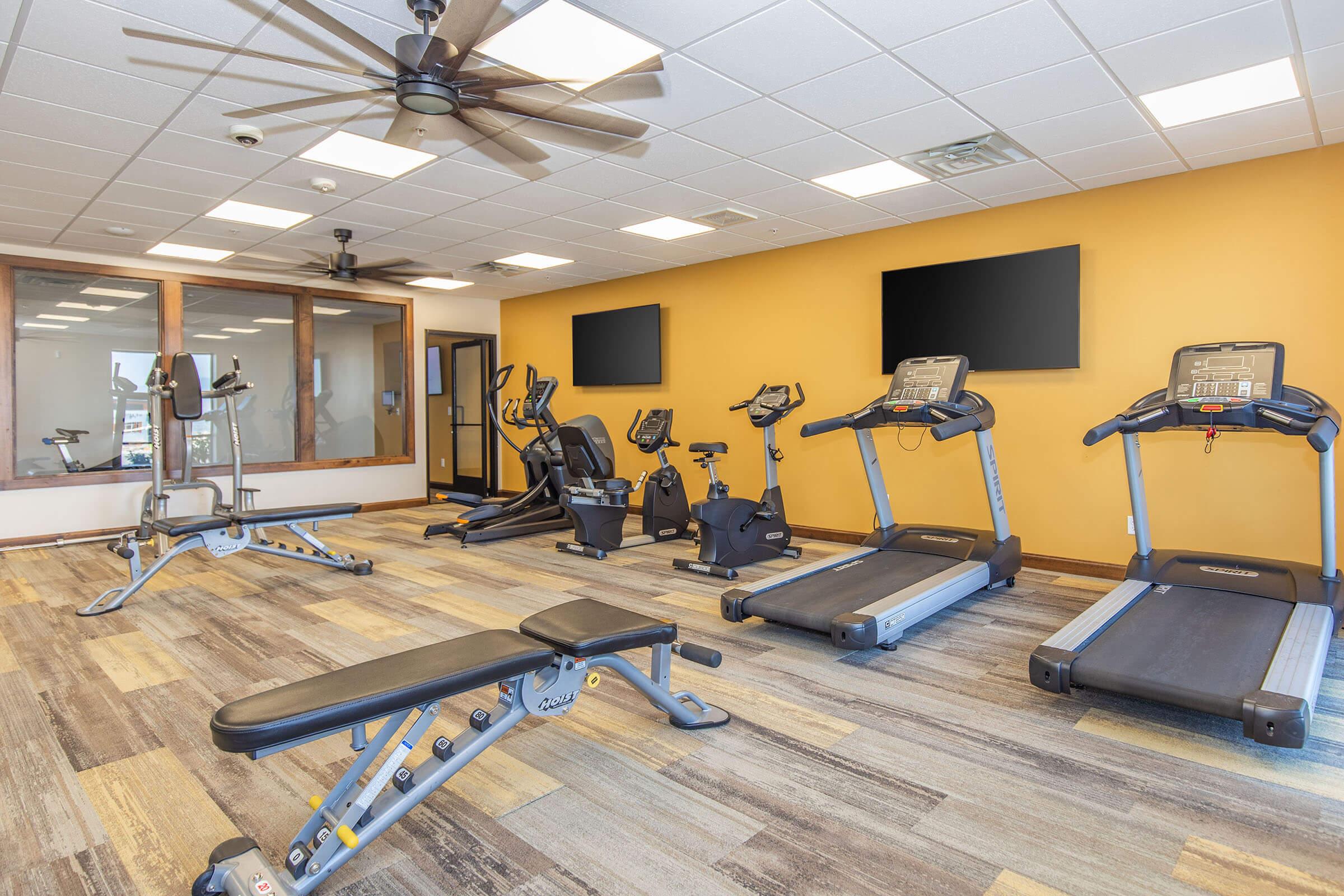 A spacious gym featuring exercise equipment including treadmills, stationary bikes, and a multi-use weight machine. The room has a modern design with wood-style flooring, ceiling fans, and large windows for natural light. Two flat-screen TVs are mounted on the wall for entertainment during workouts.