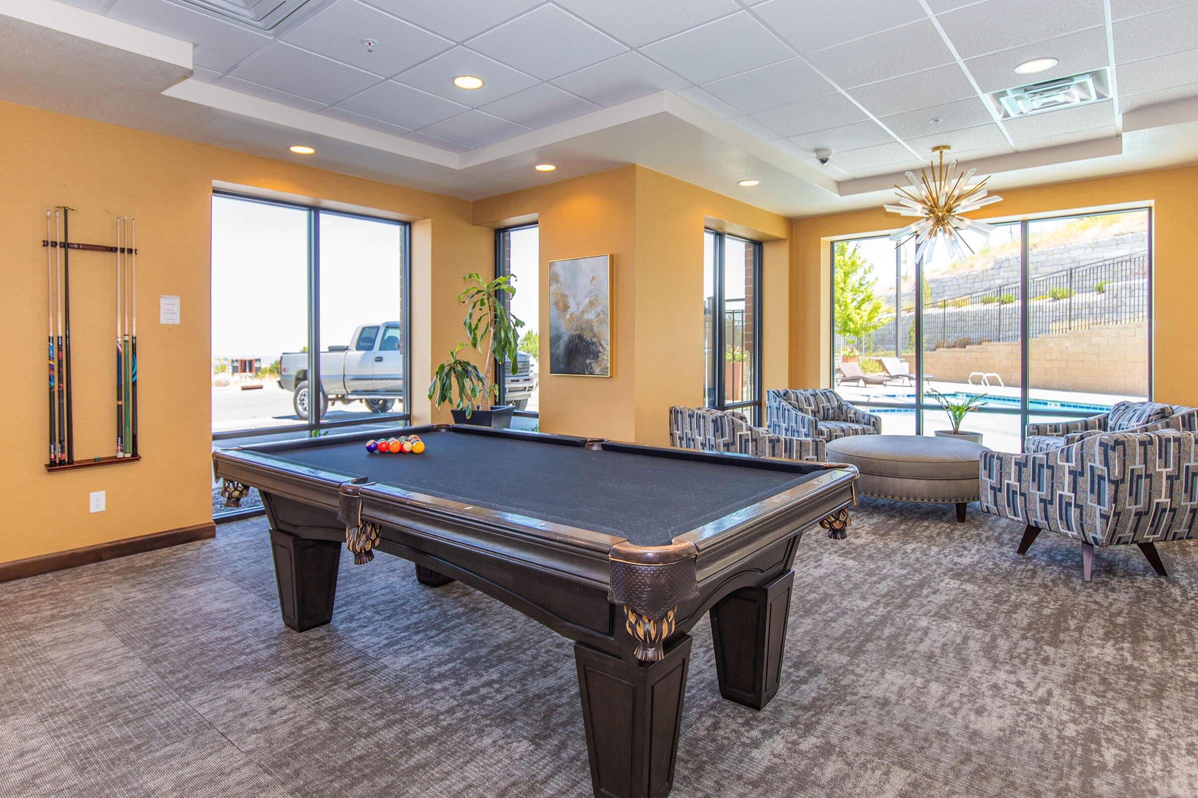 Interior view of a lounge area featuring a billiard table with colorful pool balls. There are two modern armchairs nearby and large windows providing natural light, overlooking an outdoor pool area. A plant is positioned near the windows, adding a touch of greenery to the space.