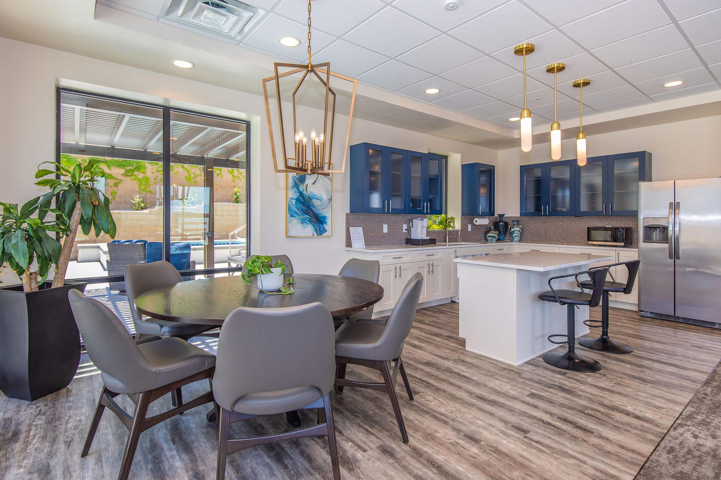 A modern kitchen featuring a round dining table with gray chairs, stylish pendant lighting, and blue cabinetry. The kitchen island includes bar stools, with stainless steel appliances and a decorative plant on the counter. Large windows provide natural light, overlooking a patio area.