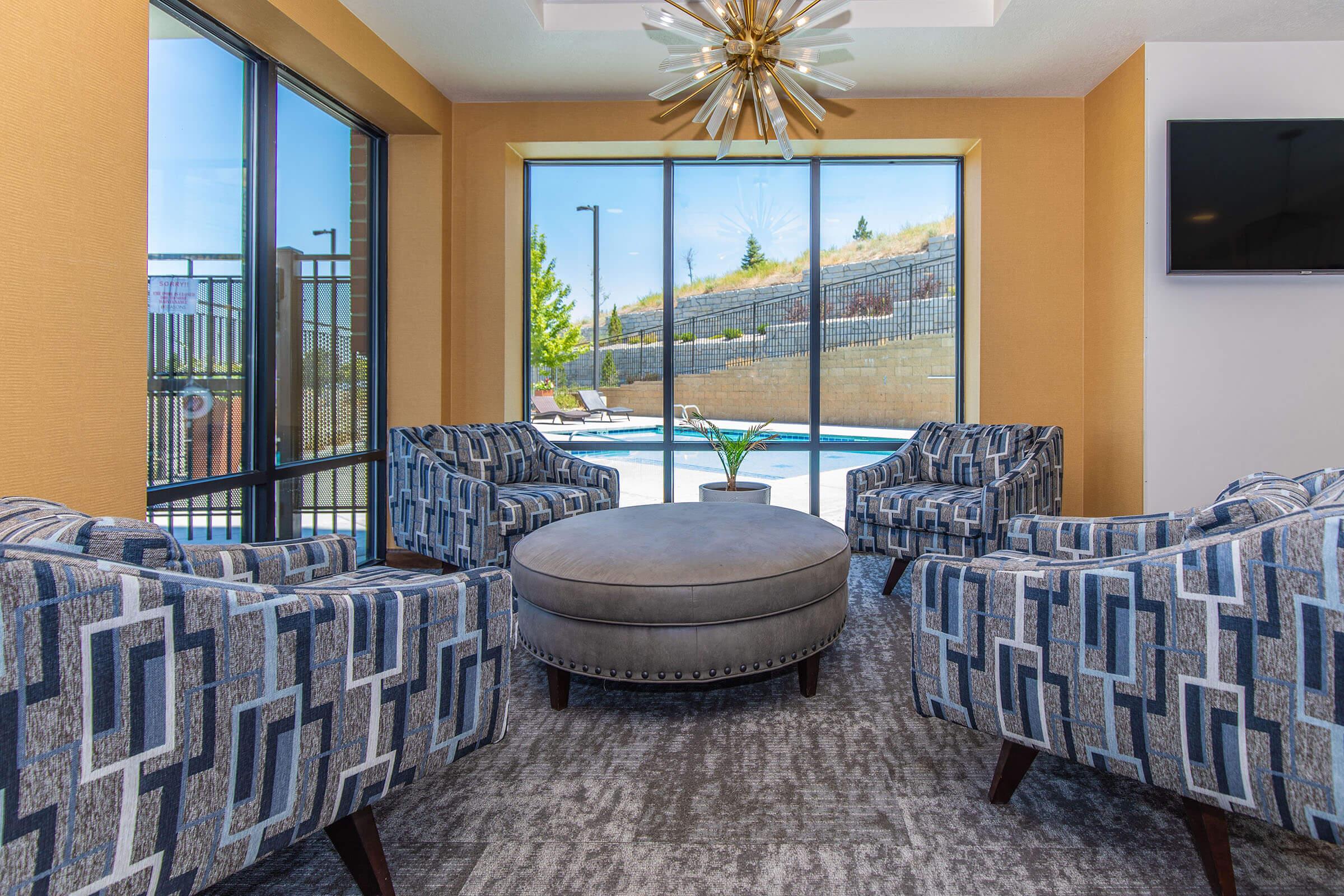 A modern lounge area featuring four patterned armchairs arranged around a round, upholstered coffee table. Large windows showcase a view of an outdoor pool area, with landscaping visible in the background. The space is bright and airy, complemented by a stylish ceiling light fixture.