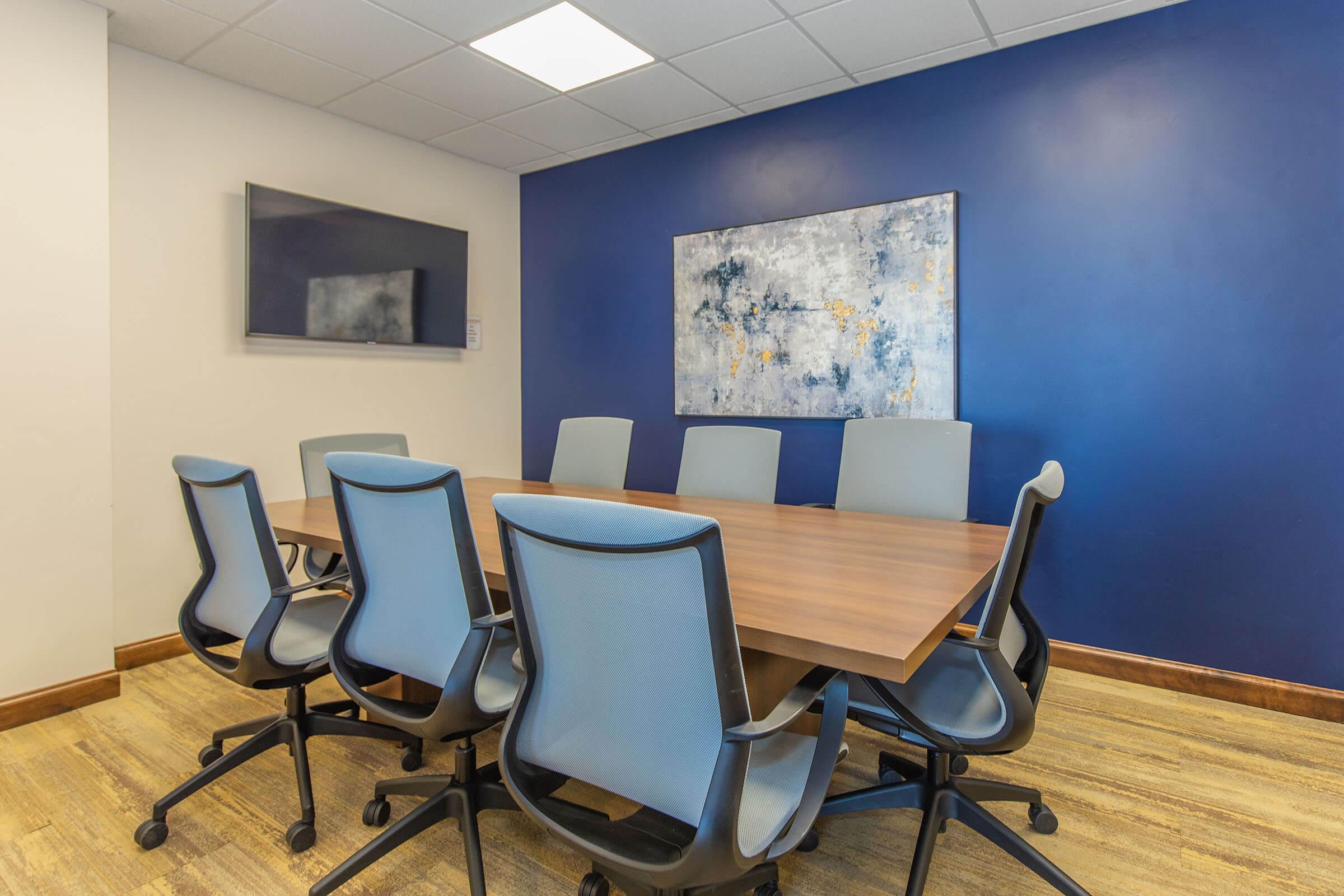 A modern conference room featuring a long wooden table surrounded by six blue office chairs. A flat-screen TV is mounted on the wall, and a large abstract painting in blue and gray tones is displayed above the table. The walls are painted navy blue, and the flooring is light wood.