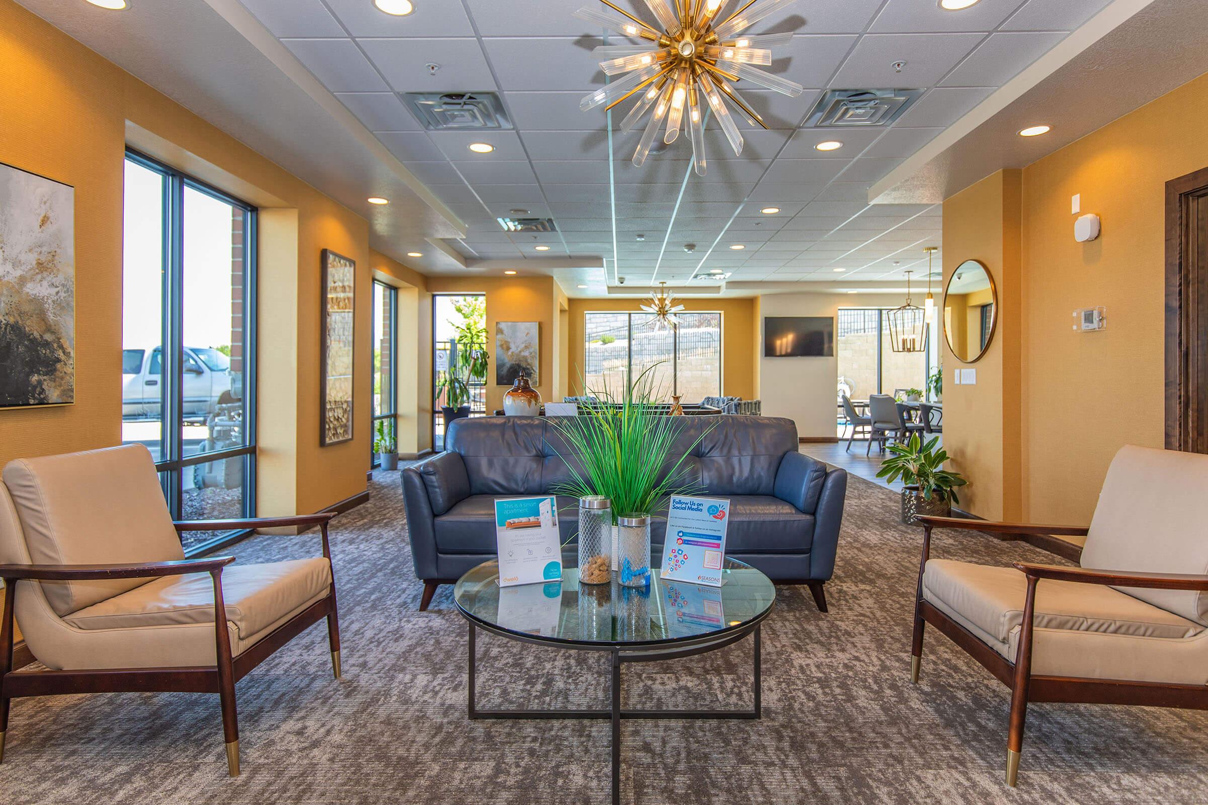 A modern lounge area featuring two beige armchairs and a leather sofa arranged around a glass coffee table. The space is adorned with plants, a decorative light fixture, and large windows offering natural light. There are informational displays on the table, and the walls are painted a warm yellow.