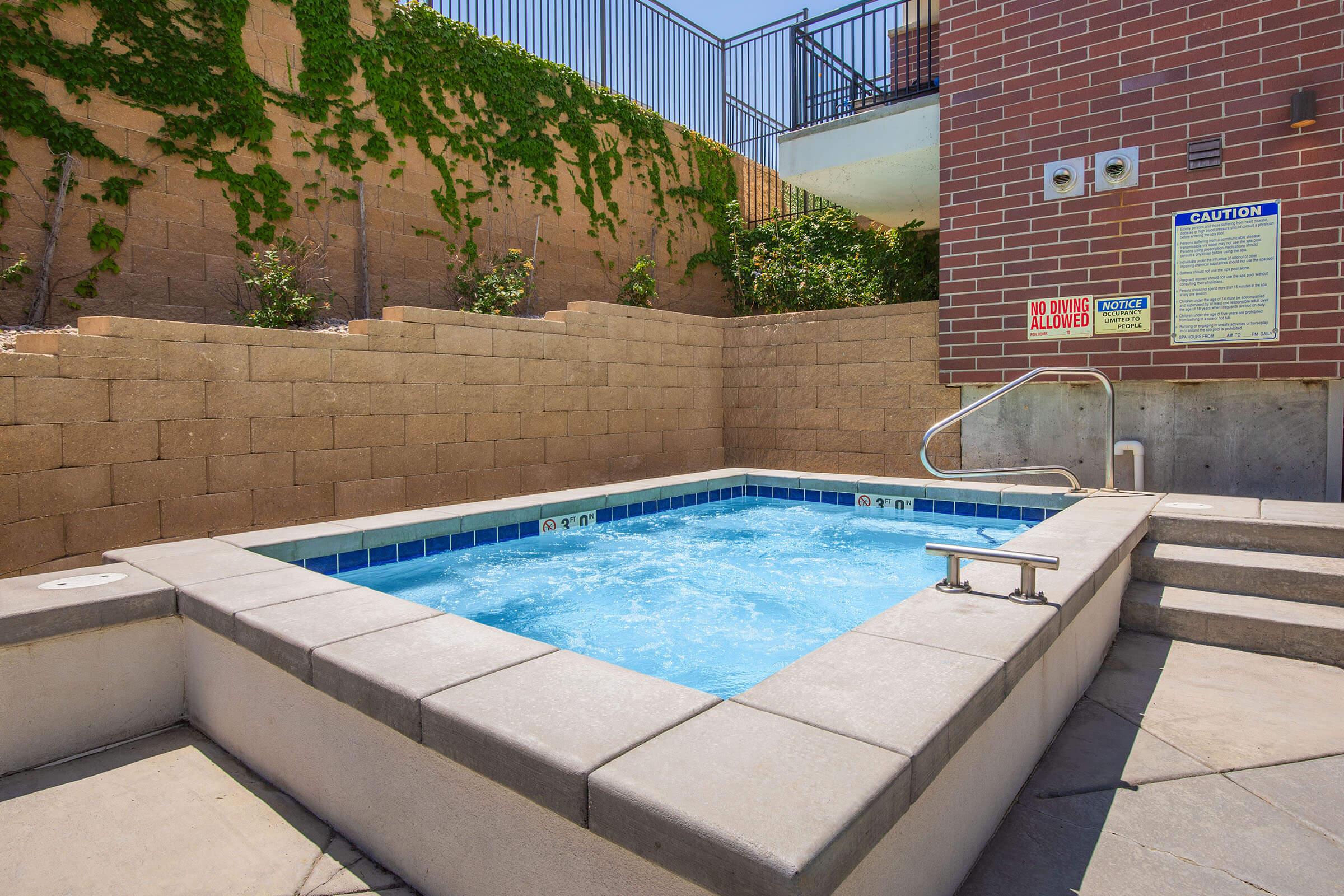 A small outdoor hot tub surrounded by a stone wall, with a blue tiled edge. The area is well-lit, featuring greenery along the walls. Safety signs are posted nearby, indicating caution and pool rules. The setting appears inviting and relaxing under clear blue skies.