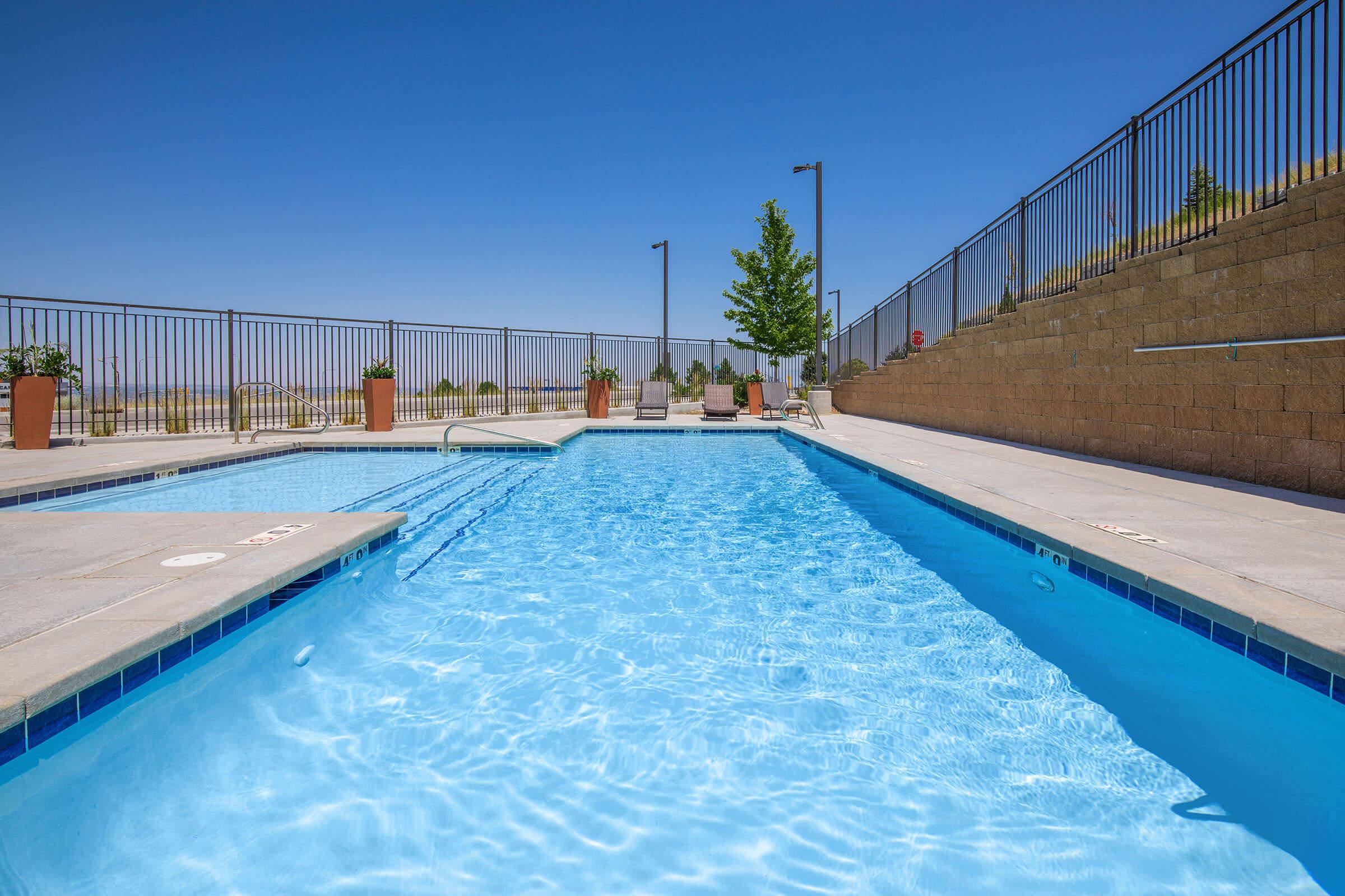 A clear blue swimming pool surrounded by a tan stone wall and a metal fence. Potted plants are placed around the pool area, and a few lounge chairs are visible in the background. The sky is bright and sunny, creating a relaxing atmosphere.