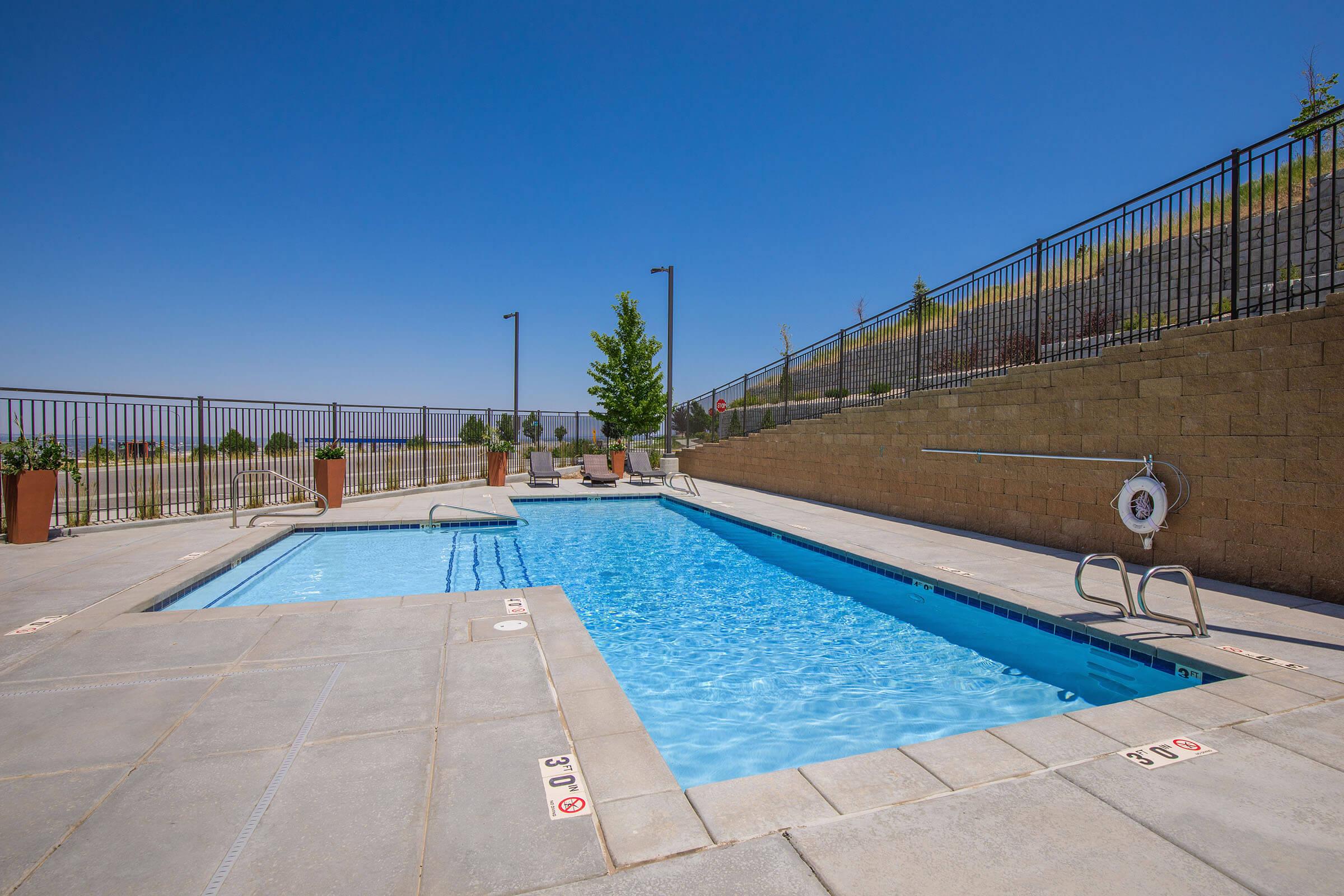 A clean and inviting swimming pool surrounded by a concrete patio, with lounge chairs nearby. Fenced perimeter with potted plants and clear blue skies overhead. The pool features steps for entry and markers for depth.