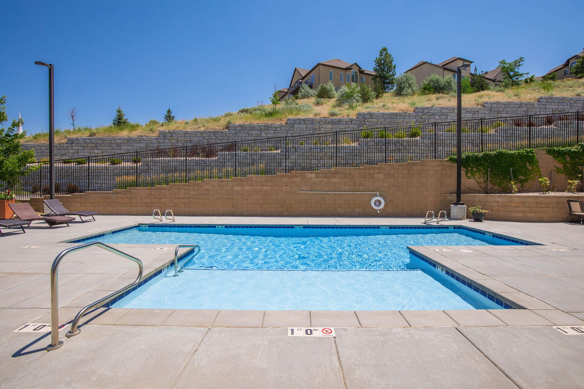 A clear blue swimming pool surrounded by a concrete deck. The pool features a ladder and is bordered by lounge chairs. In the background, there are landscaped hills and residential buildings. Bright sunlight casts shadows, creating a relaxing outdoor atmosphere.