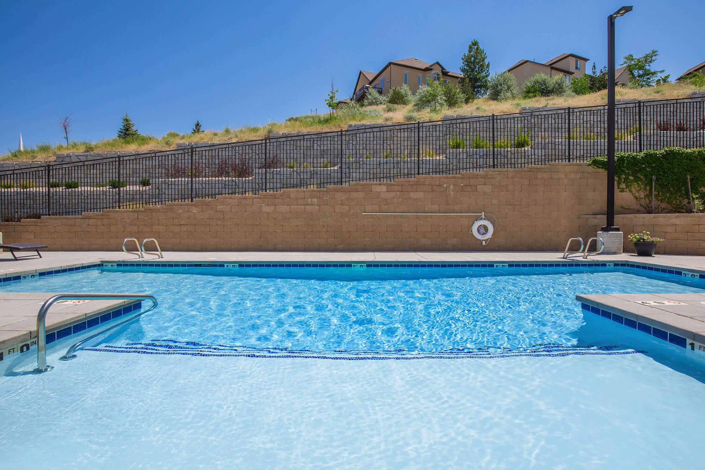 A bright blue swimming pool with clear water, surrounded by a stone patio. In the background, there are grassy hills and residential buildings. The setting is sunny, creating a serene atmosphere. A life preserver is mounted on the wall near the pool.