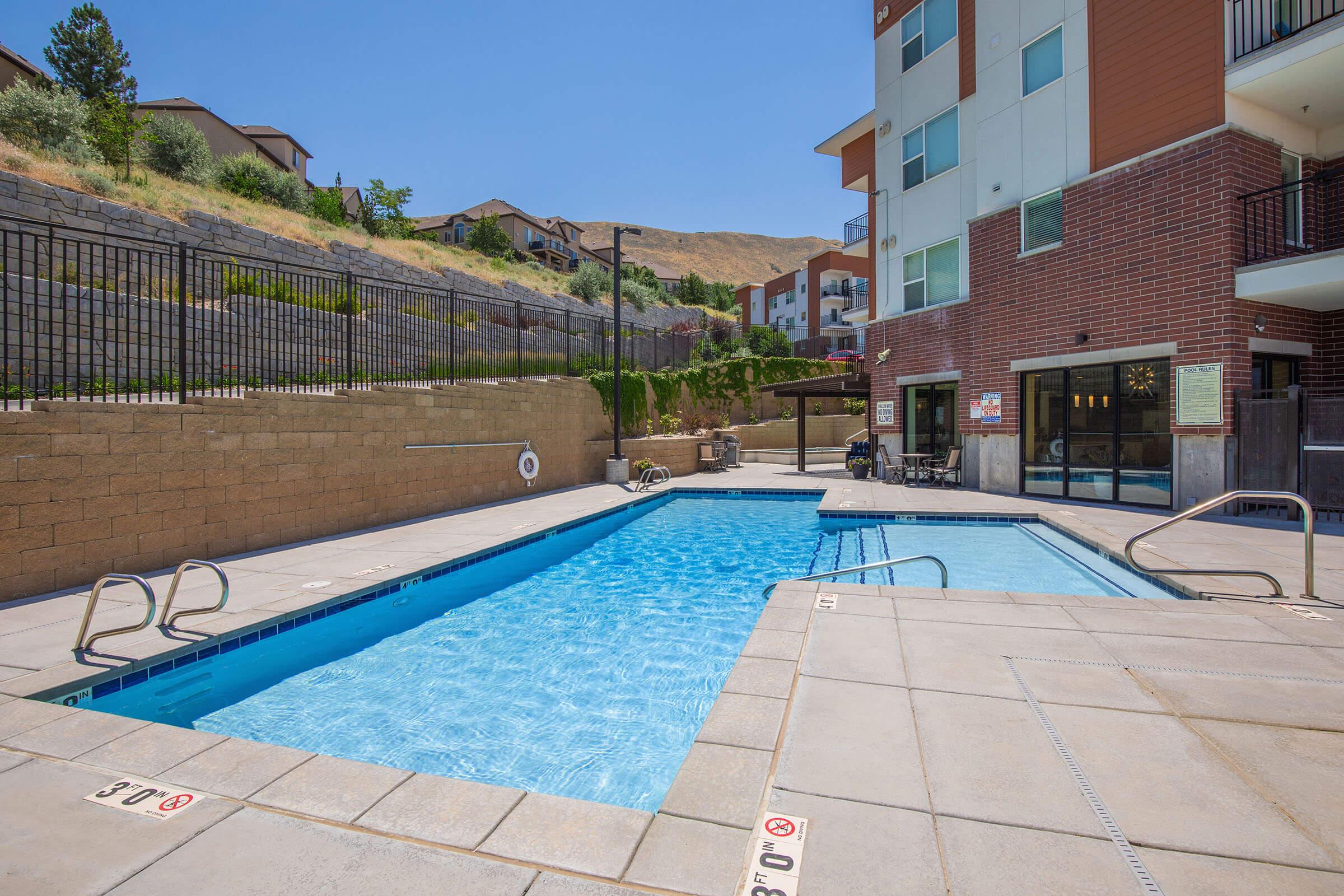 A clear view of a swimming pool surrounded by a tiled deck, with lounge chairs arranged nearby. The pool area features a safety railing and is bordered by a residential building and landscaped hills. The sky is sunny and blue, adding to the inviting atmosphere of the outdoor space.