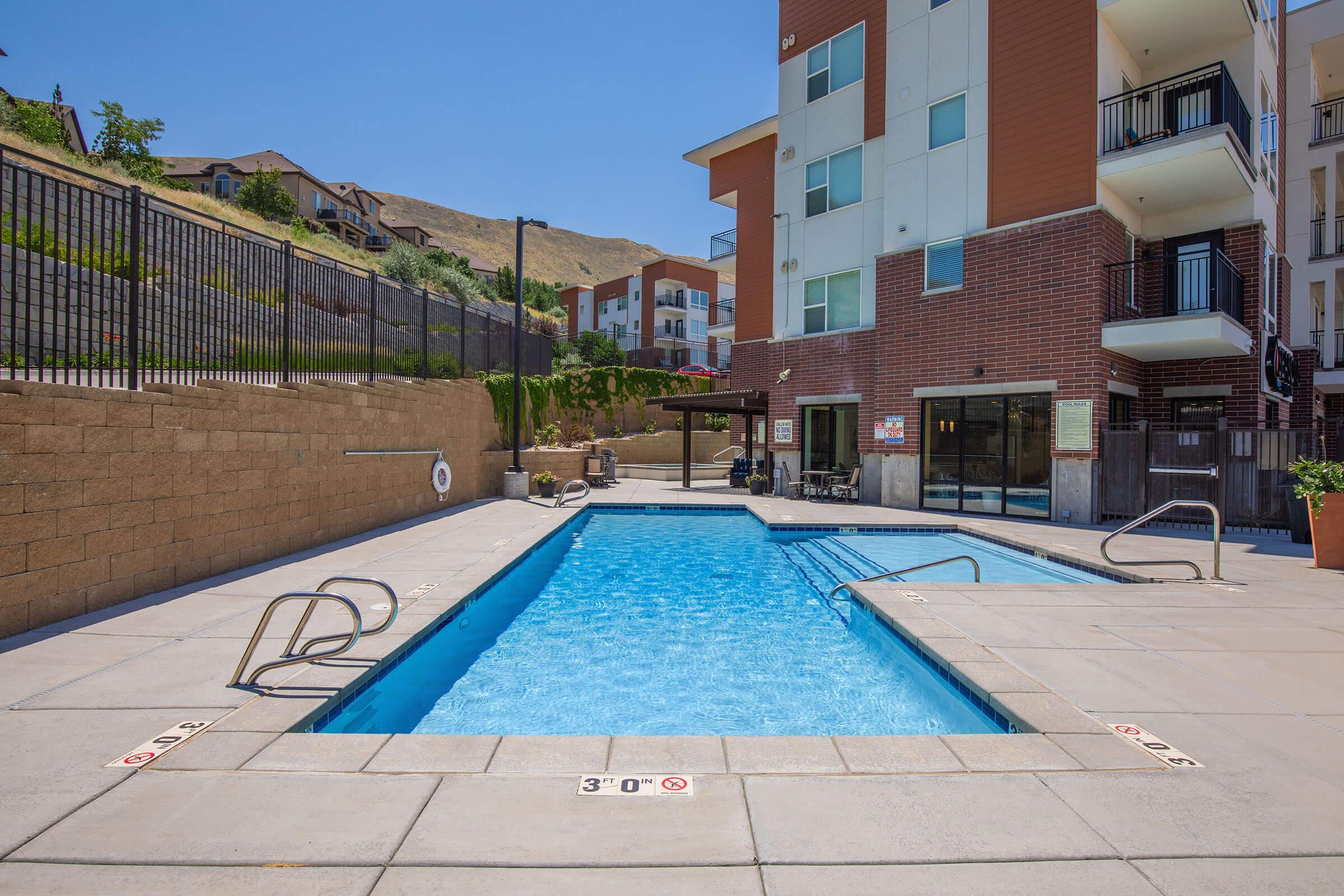 A swimming pool on a sunny day, featuring clear blue water, steps, and safety markings. Surrounded by a landscaped area with a few residential buildings in the background, the scene conveys a relaxing outdoor atmosphere perfect for leisure and recreation.