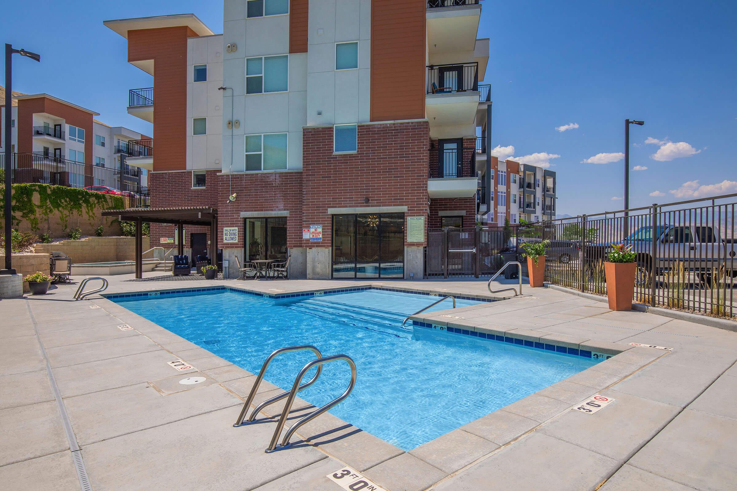 A bright outdoor swimming pool surrounded by a concrete deck, with pool ladders on either side. In the background, a modern apartment building with multiple floors and balconies is visible. Potted plants add a touch of greenery, and the sky is clear with a few clouds.