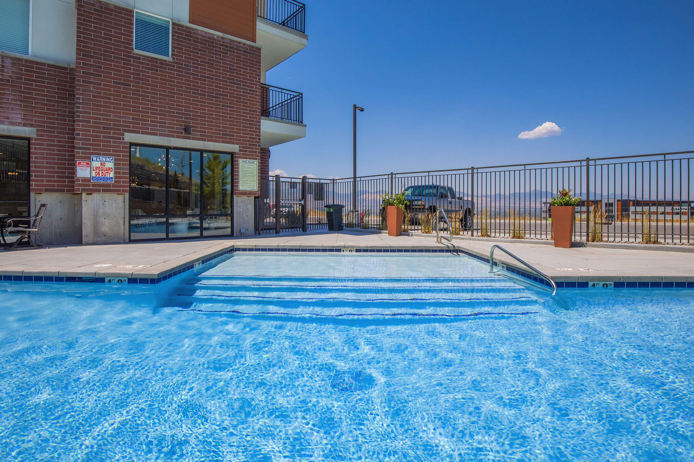 A clear, inviting swimming pool with steps leading into it, situated near a modern building. The pool area features a fenced perimeter, and behind it is a scenic view of mountains and blue sky. The sunlight reflects off the water, creating a serene atmosphere.