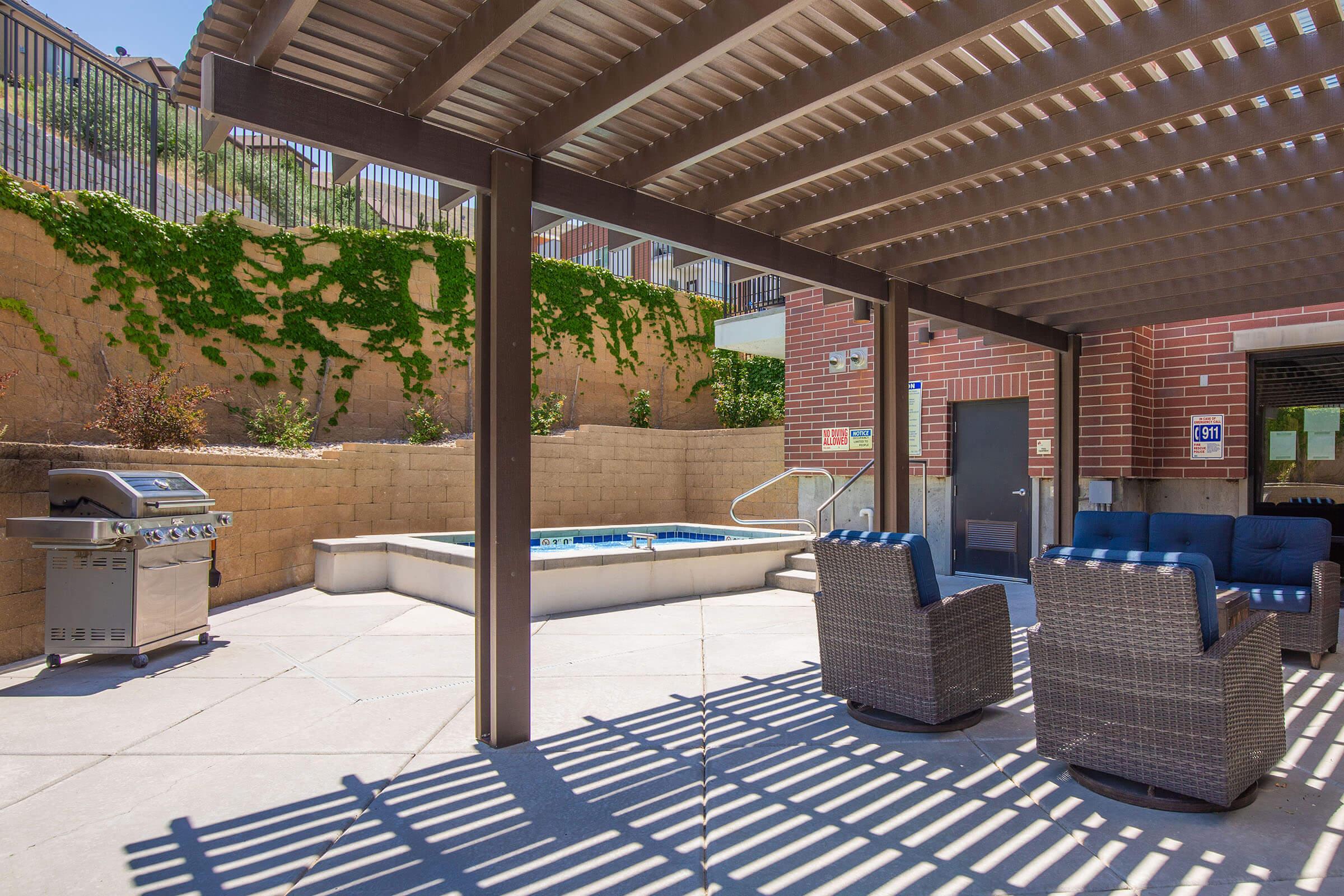 Outdoor patio area featuring a shaded seating arrangement with two wicker chairs and a barbecue grill. A small hot tub is visible in the background, surrounded by a stone wall and greenery. The area is well-lit, showcasing a clean and inviting atmosphere for relaxation and outdoor cooking.