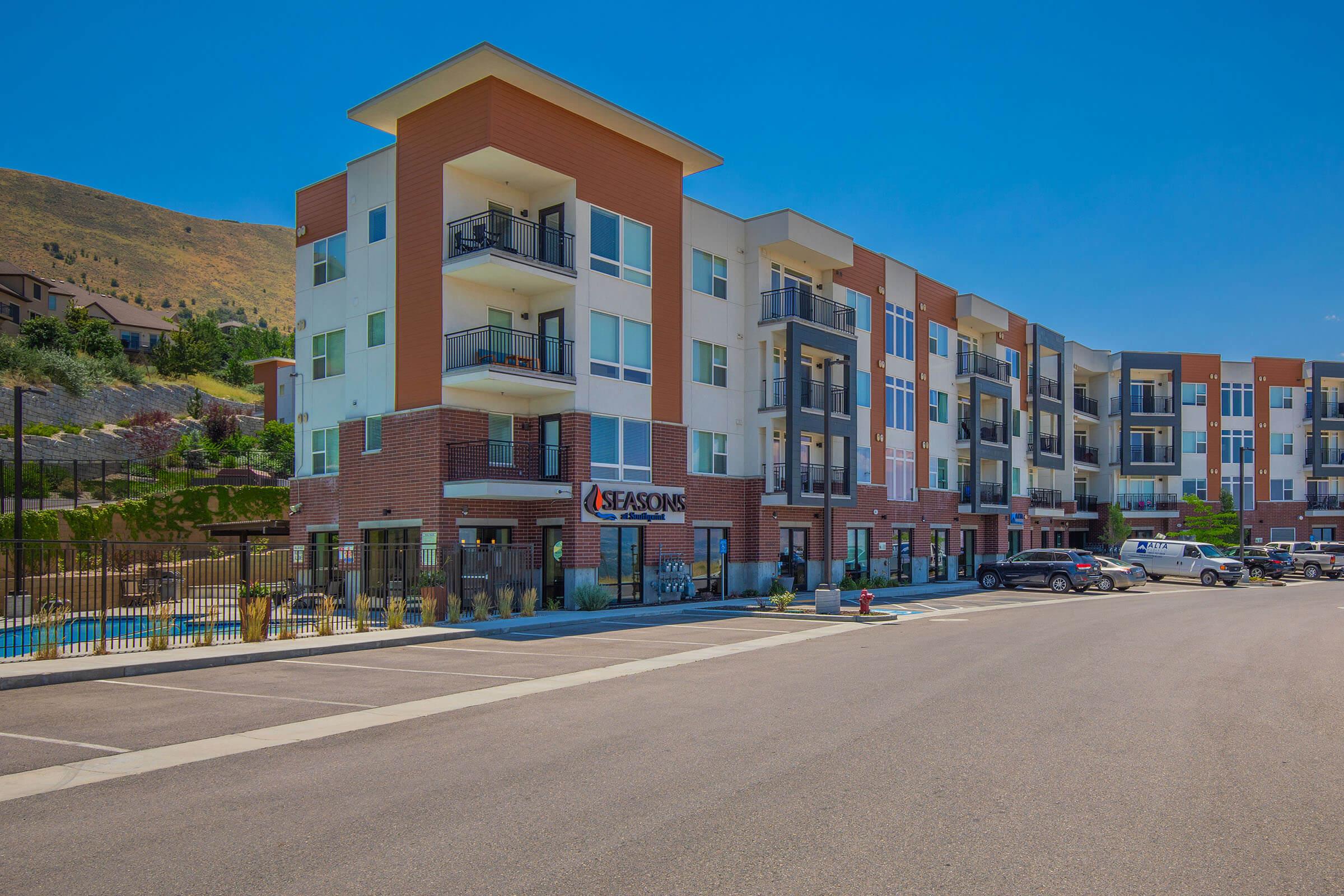 A multi-story apartment building named "Seasons" featuring modern architecture, balconies, and a parking area. In the foreground, there is a wide parking lot with several parked cars, and in the background, green hills under a clear blue sky. A swimming pool is visible beside the building, surrounded by a fence.