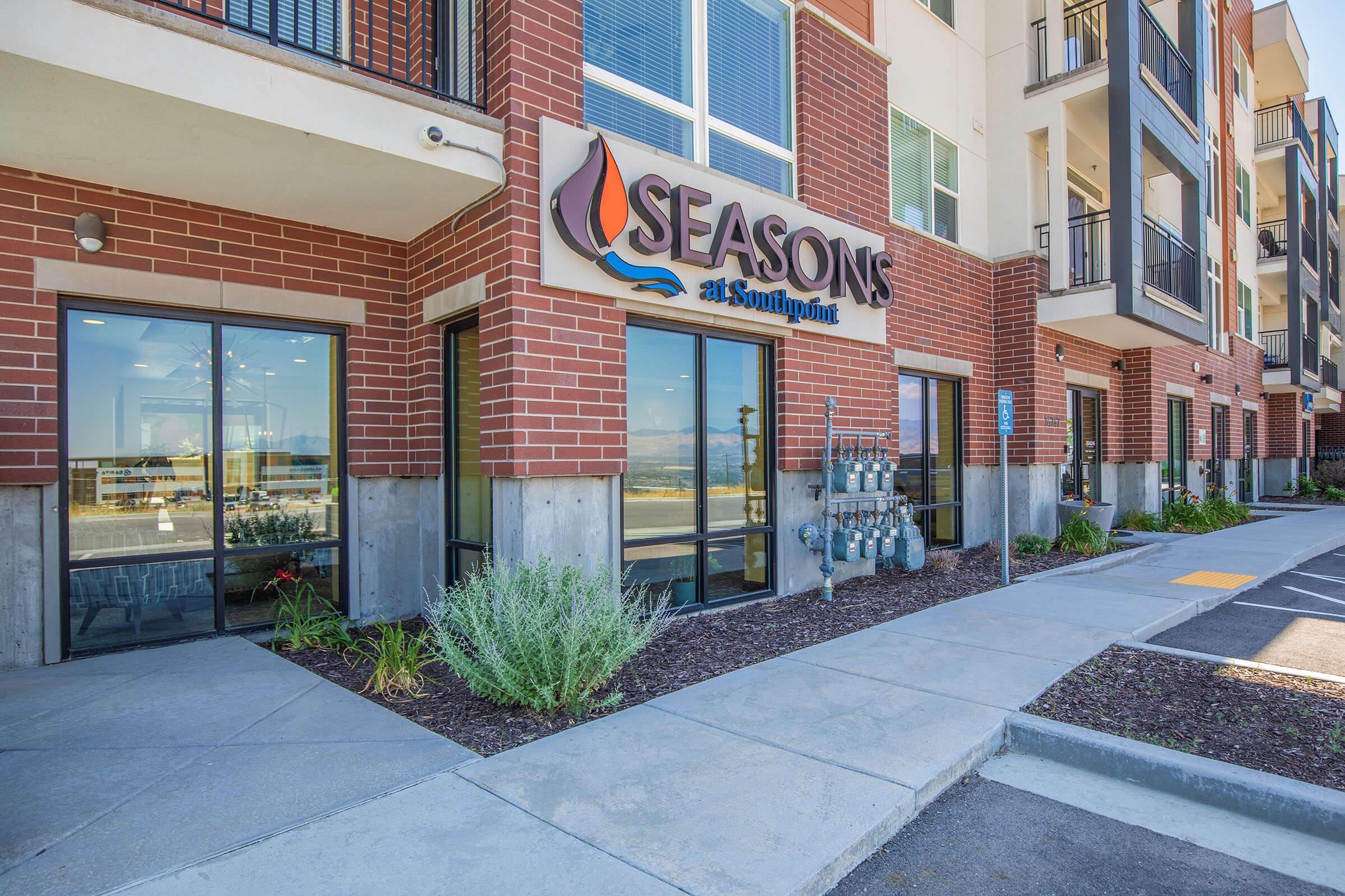 Exterior view of an apartment building named "Seasons at Southpoint." The entrance features large glass windows and a prominent sign. The walkway is paved, with landscaped greenery and utility meters visible nearby. A parking area is present in the front, along with a clear blue sky in the background.