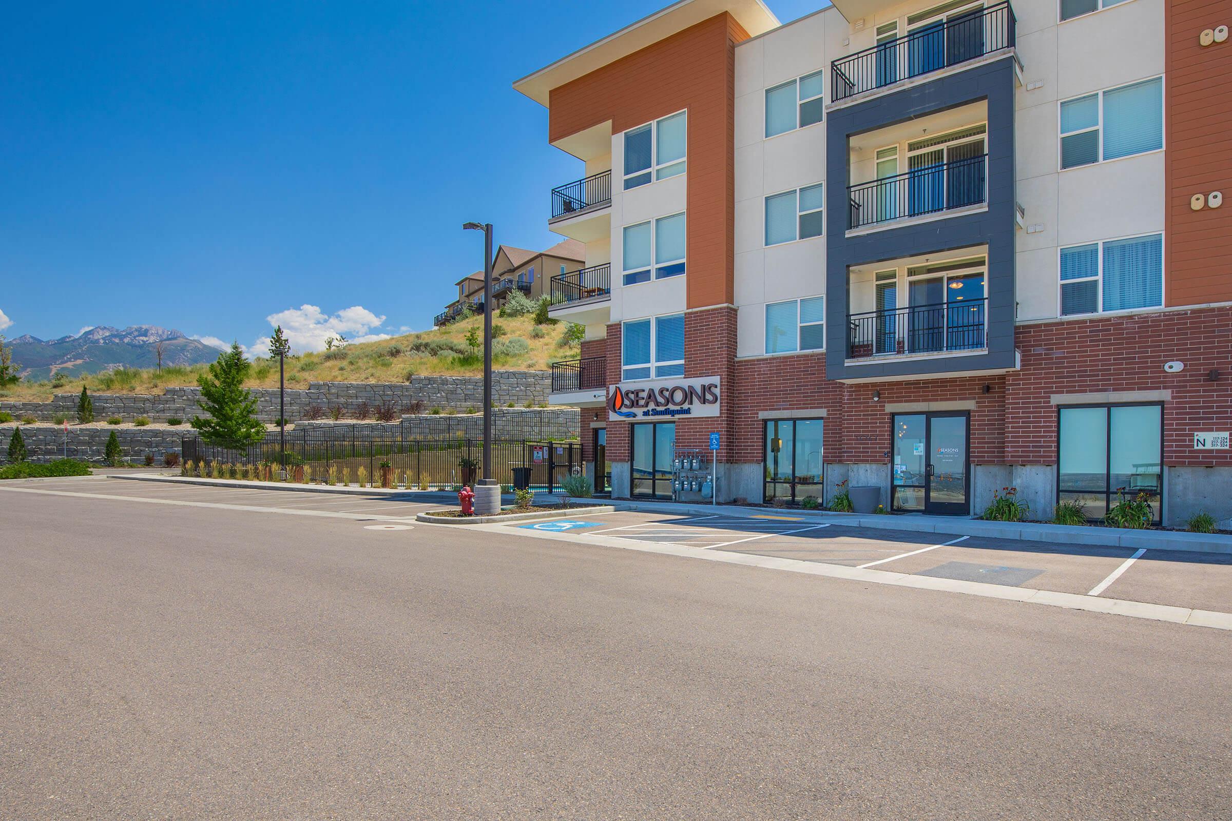 A modern multi-story apartment building named "Seasons" located in a scenic area. The building features a mix of brick and cream-colored facade, balconies on upper levels, and parking spaces in front. The background showcases mountains under a clear blue sky.