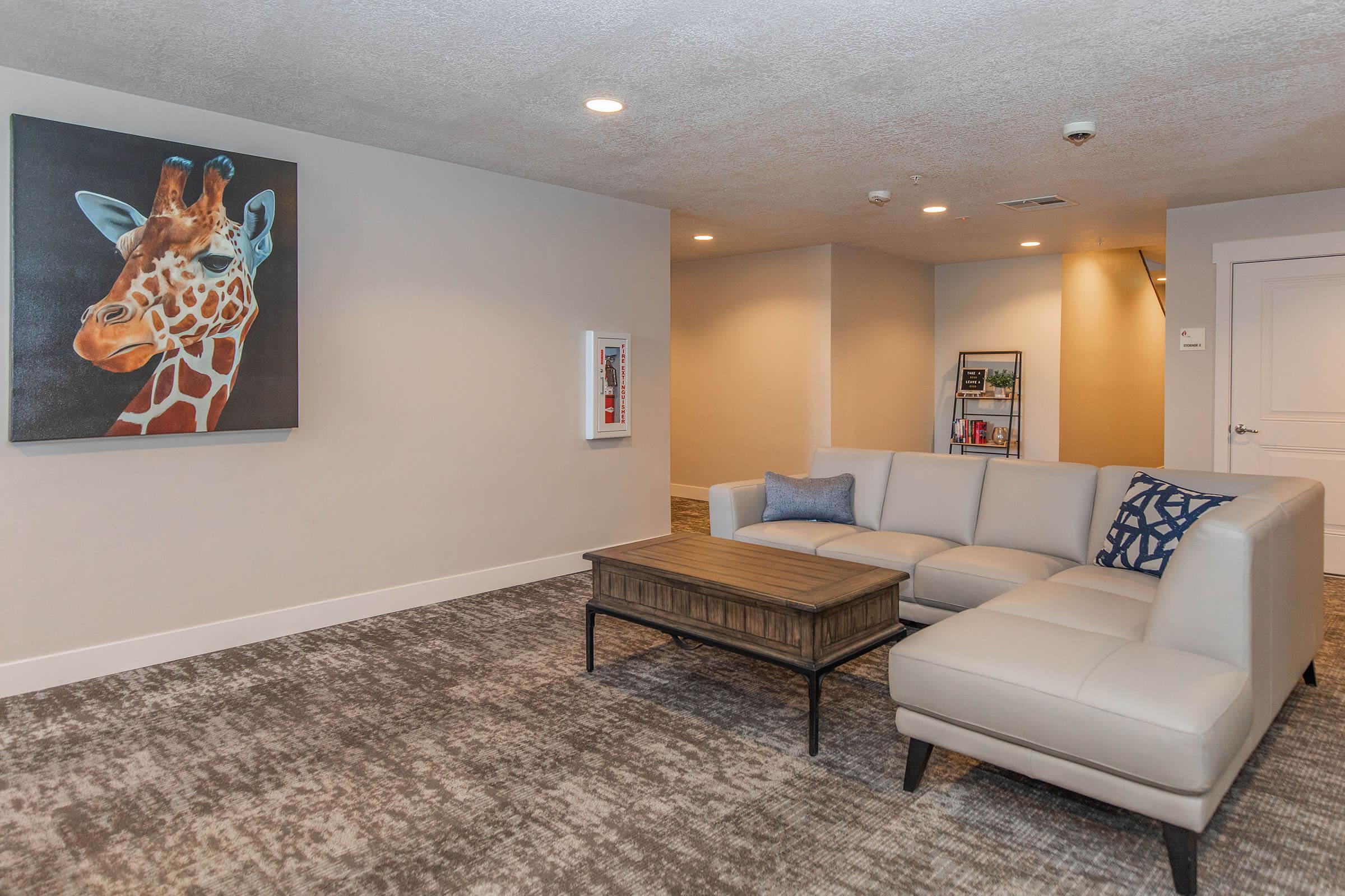 A modern, stylish lounge area featuring a light-colored sectional sofa with decorative cushions. There is a wooden coffee table in the center. A colorful painting of a giraffe hangs on the wall, and a small bookshelf is visible in the background along with warm lighting and neutral-colored walls.