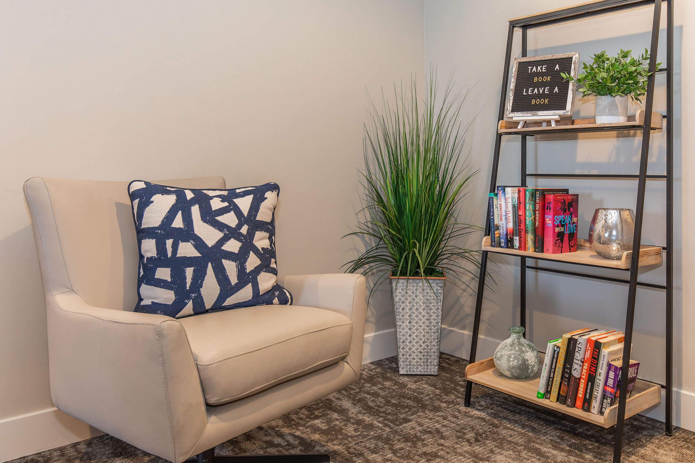 A cozy reading nook featuring a light-colored armchair with a blue geometric patterned pillow. Nearby, a tall plant in a decorative pot stands next to a modern shelving unit with books, a small glass vase, a candle, and a sign that reads "TAKE A BOOK, LEAVE A BOOK." The floor has a soft gray carpet.