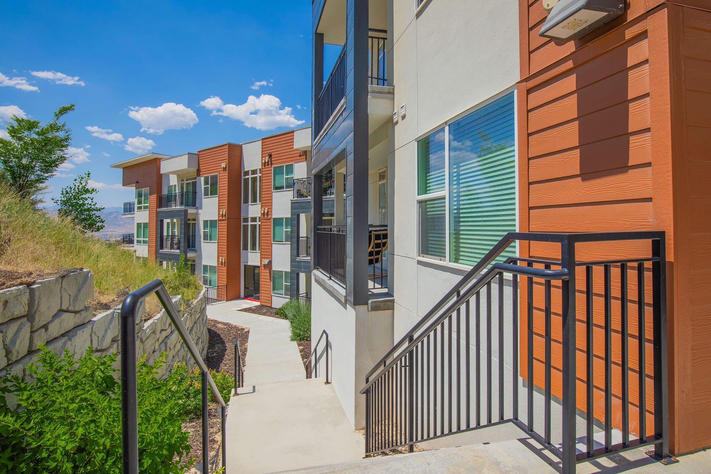 A pathway leading down between modern apartment buildings with a mix of brown and cream siding. Lush greenery flanks the stairway, and a clear blue sky with scattered clouds is visible overhead. The scene is bright and inviting, showcasing a well-maintained residential area.