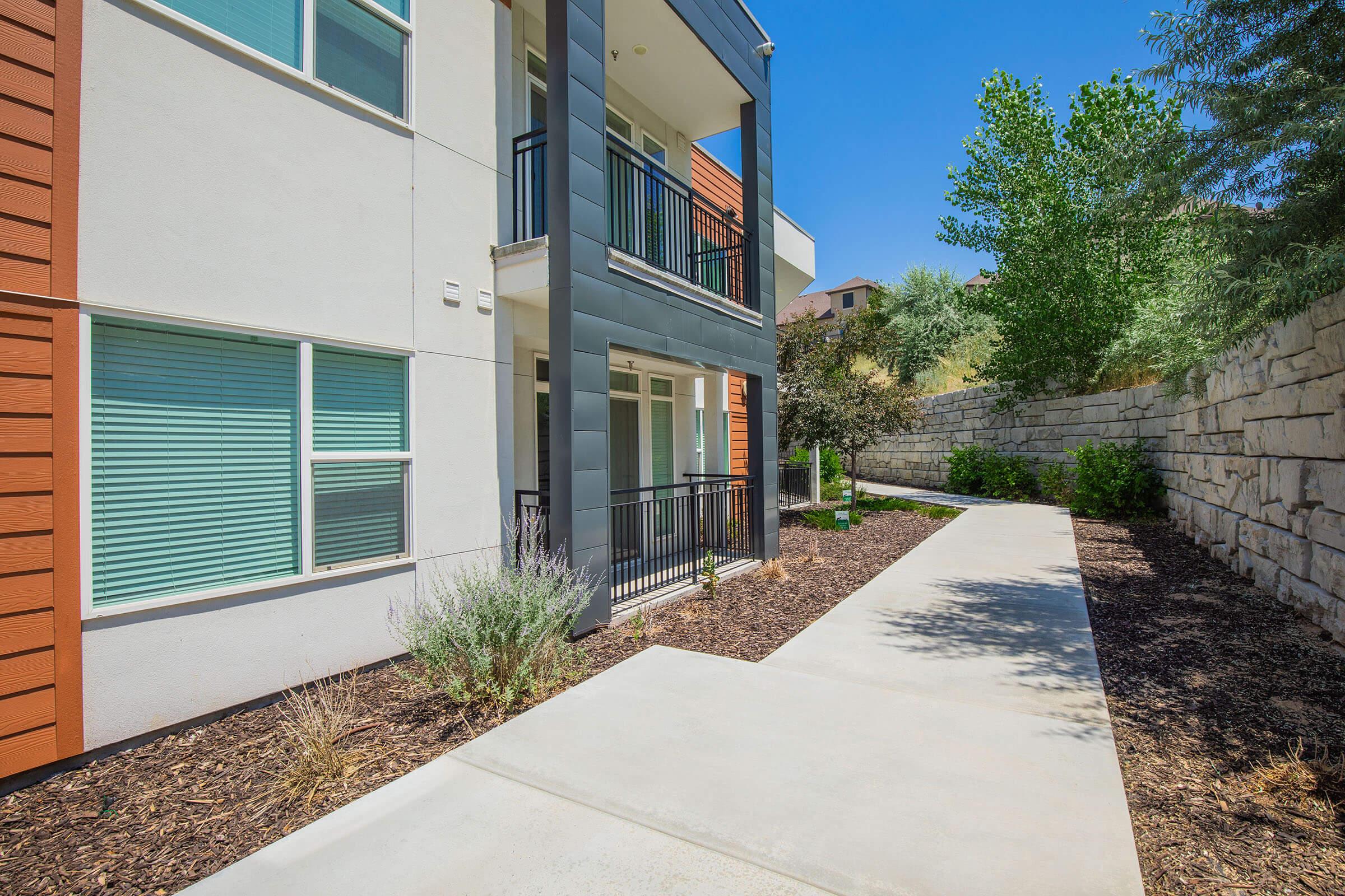 A bright, modern residential building featuring a landscaped path leading to the entrance, bordered by plants and trees. The exterior walls are a mix of light and dark materials, with large windows allowing natural light. The surrounding area includes a stone wall and greenery, enhancing the inviting atmosphere.