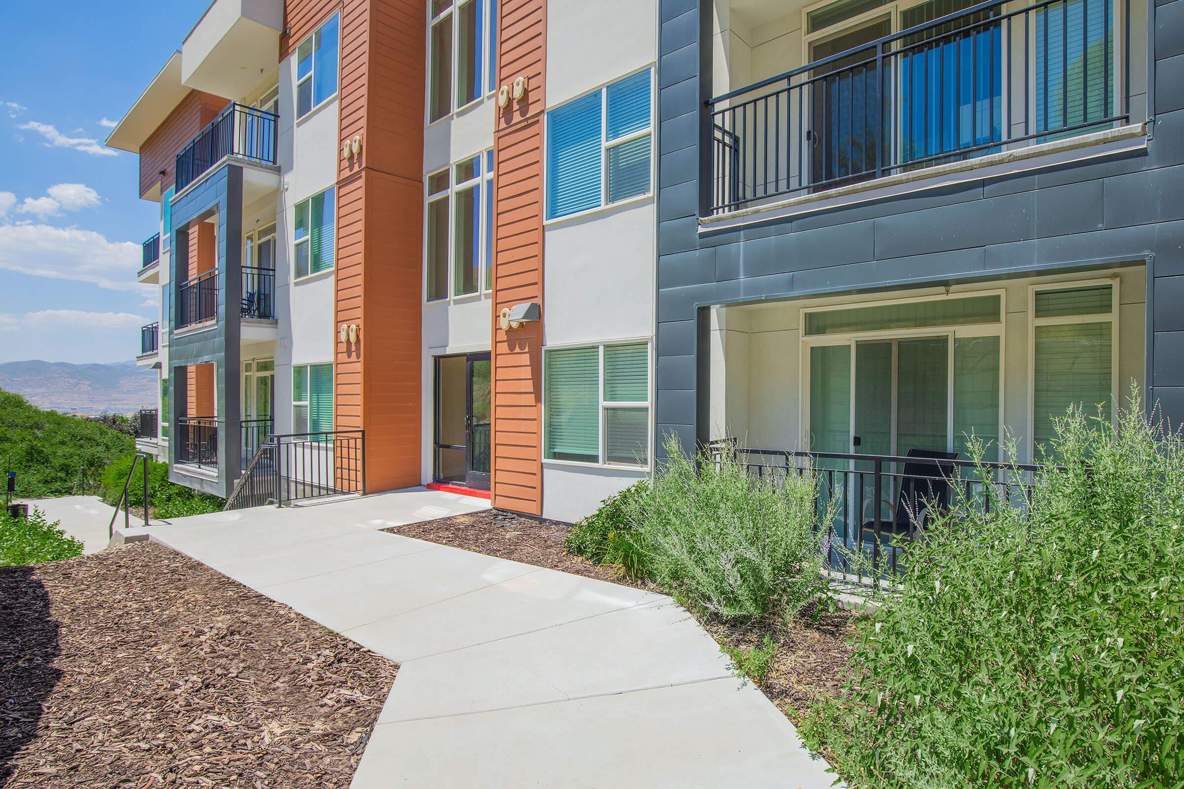 A modern apartment community featuring multiple stories with a combination of orange and gray exterior finishes. The walkway leads to the entrance, surrounded by greenery and shrubs. There are balconies on the upper floors, with large windows allowing natural light to illuminate the interiors.