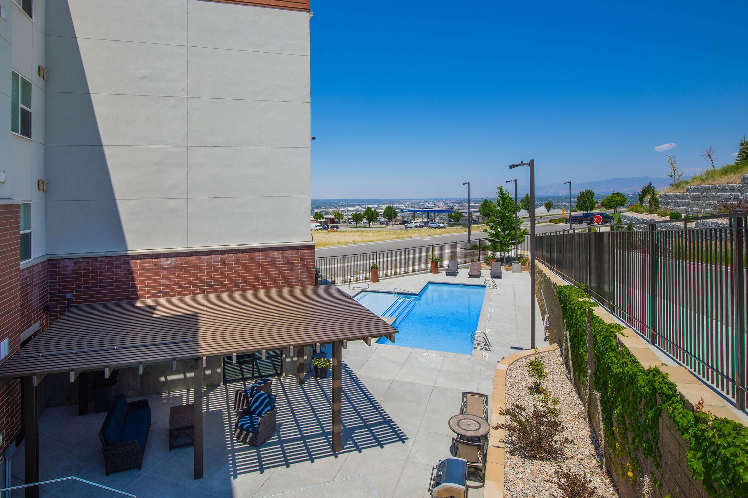 A view of an outdoor swimming pool surrounded by a patio area with seating and shade. The pool is situated next to a building, with landscaped greenery and a fence in the background. The scene is set against a clear blue sky, showcasing a distant cityscape and mountains in the background.